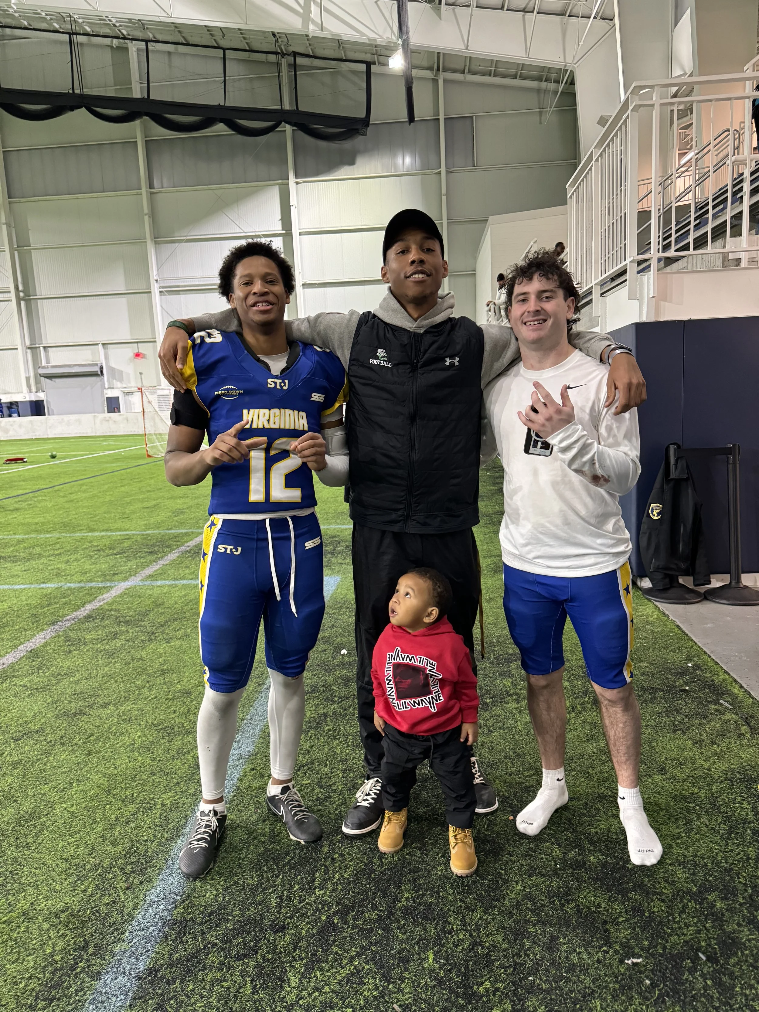 Four young men and a small child posing together inside an indoor sports facility, with the young men wearing athletic clothing, one in a Virginia football uniform.