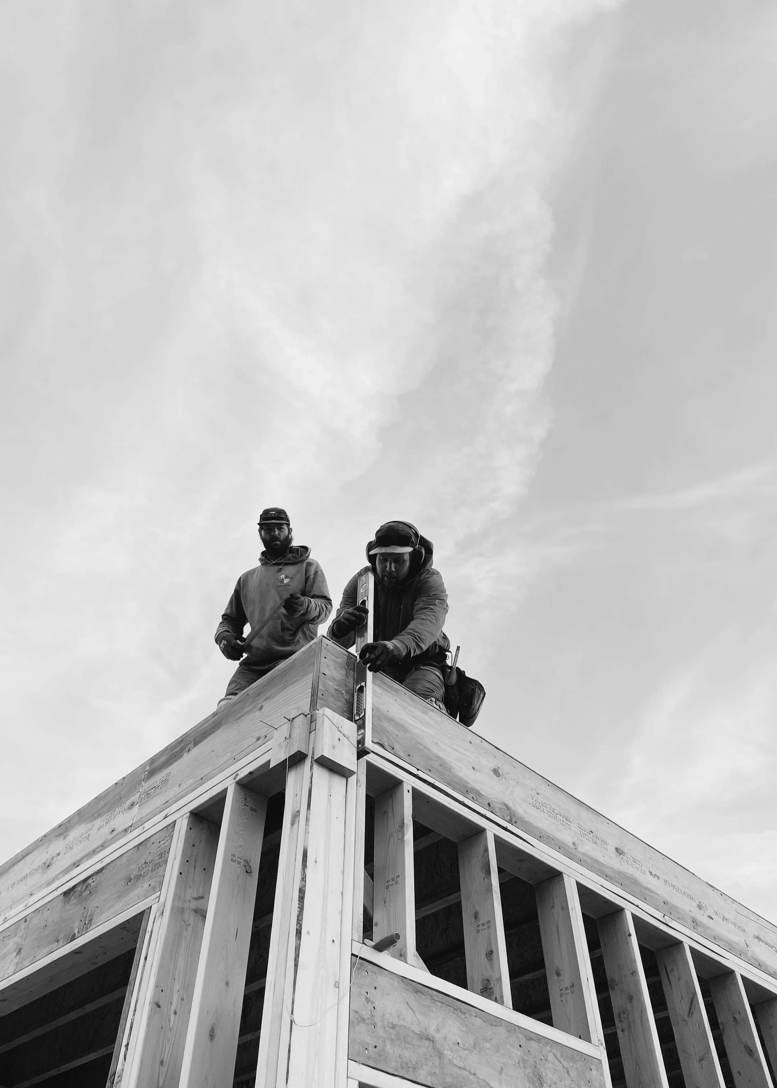 Two construction workers on top of a building under construction, with a cloudy sky in the background. One worker is wearing headphones and the other is standing behind him.
