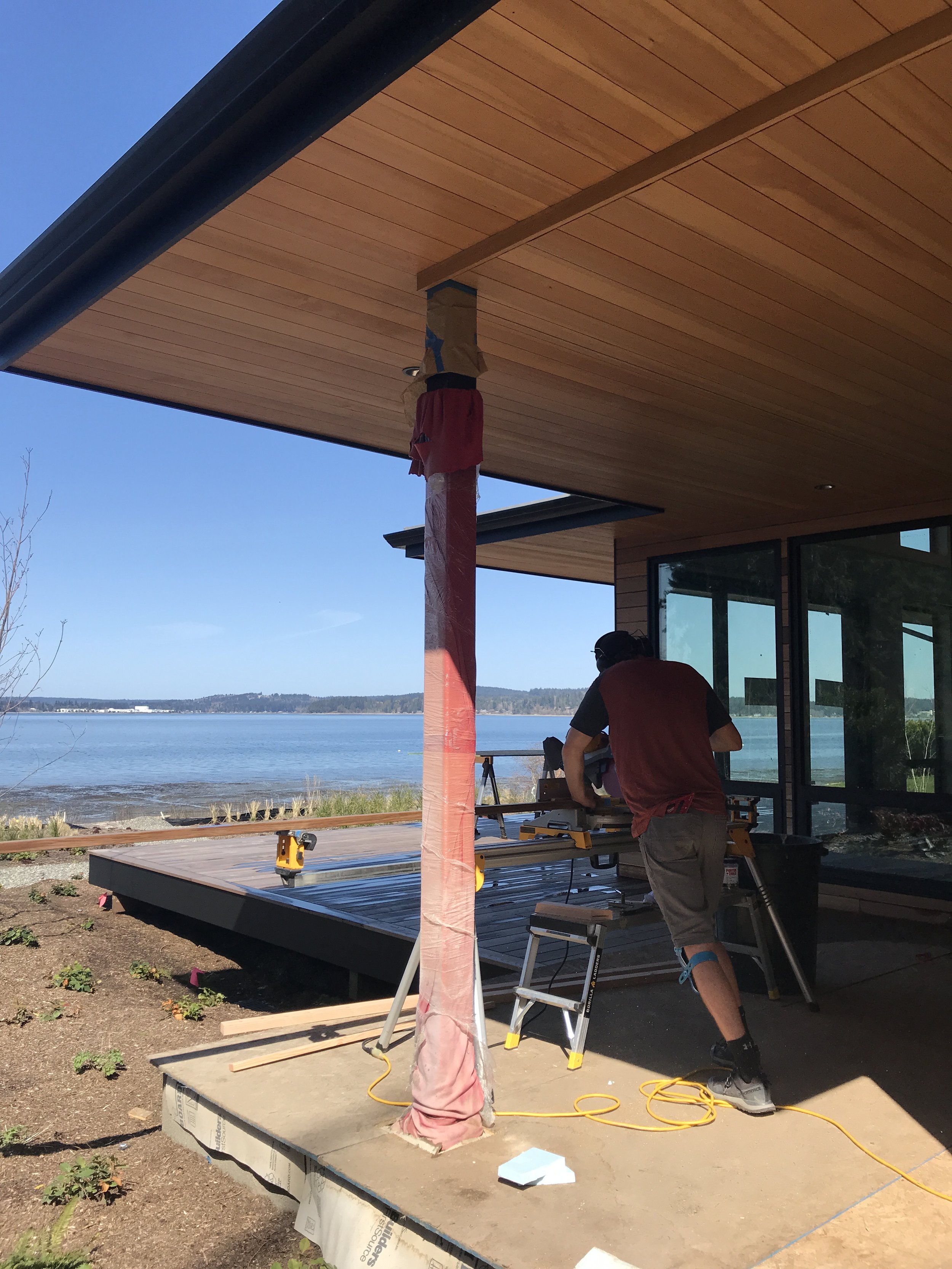 Construction worker installing a house support post on a wooden deck near a body of water, with the house featuring large glass windows and a wooden roof extension.
