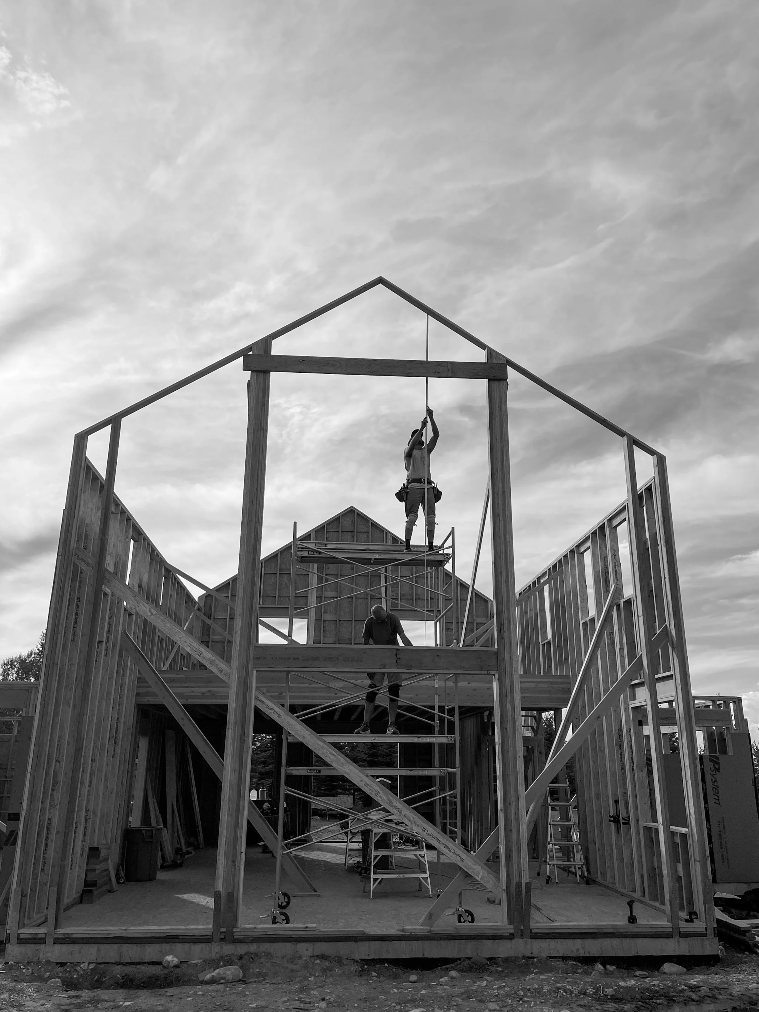 Construction workers building a wooden house framework, with scaffolding and tools, under a cloudy sky.