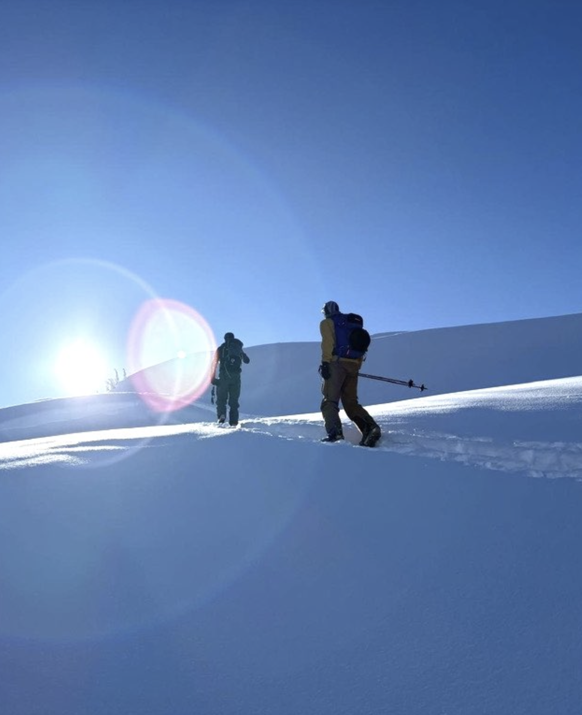 Two backcountry skiers testing product on snow-covered terrain in Squamish under a bright blue sky with sunlight and lens flare.