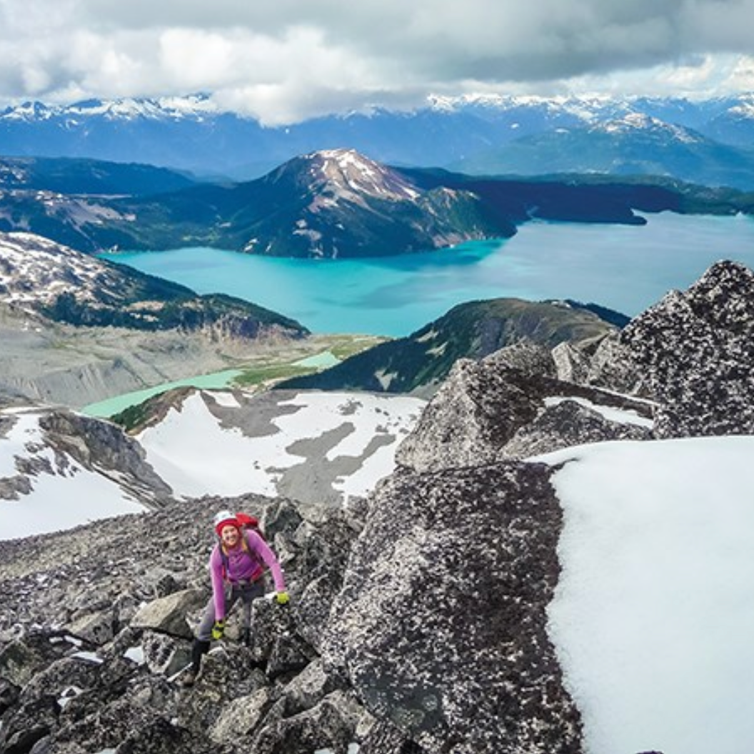 A person in a pink jacket and red hat climbing on rocky mountain terrain in Squamish.