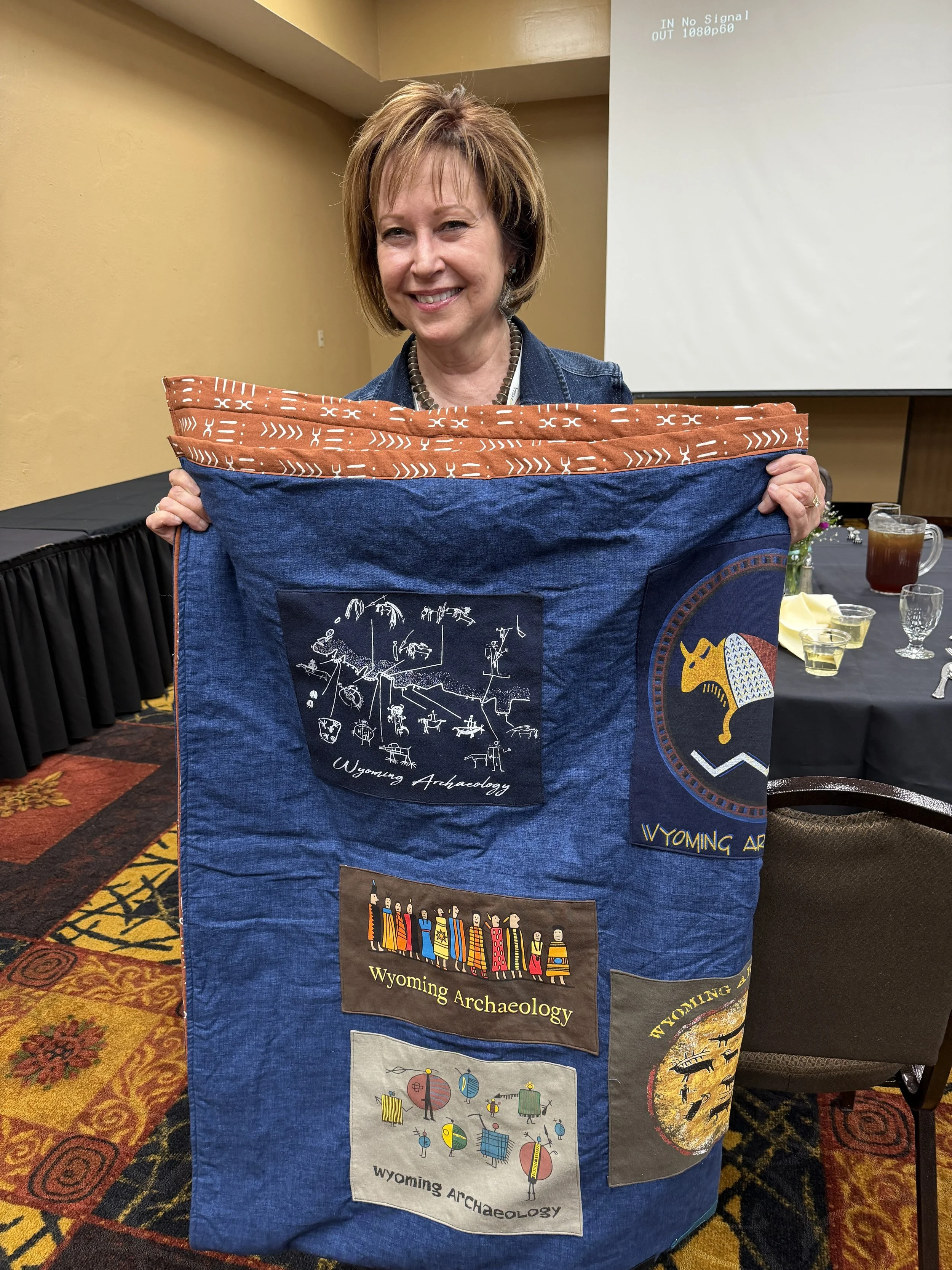 Judy Wolf showing off her score from the silent auction: a quilt made by Becca Mashak's mother featuring old Wyoming Archaeology t-shirts. 