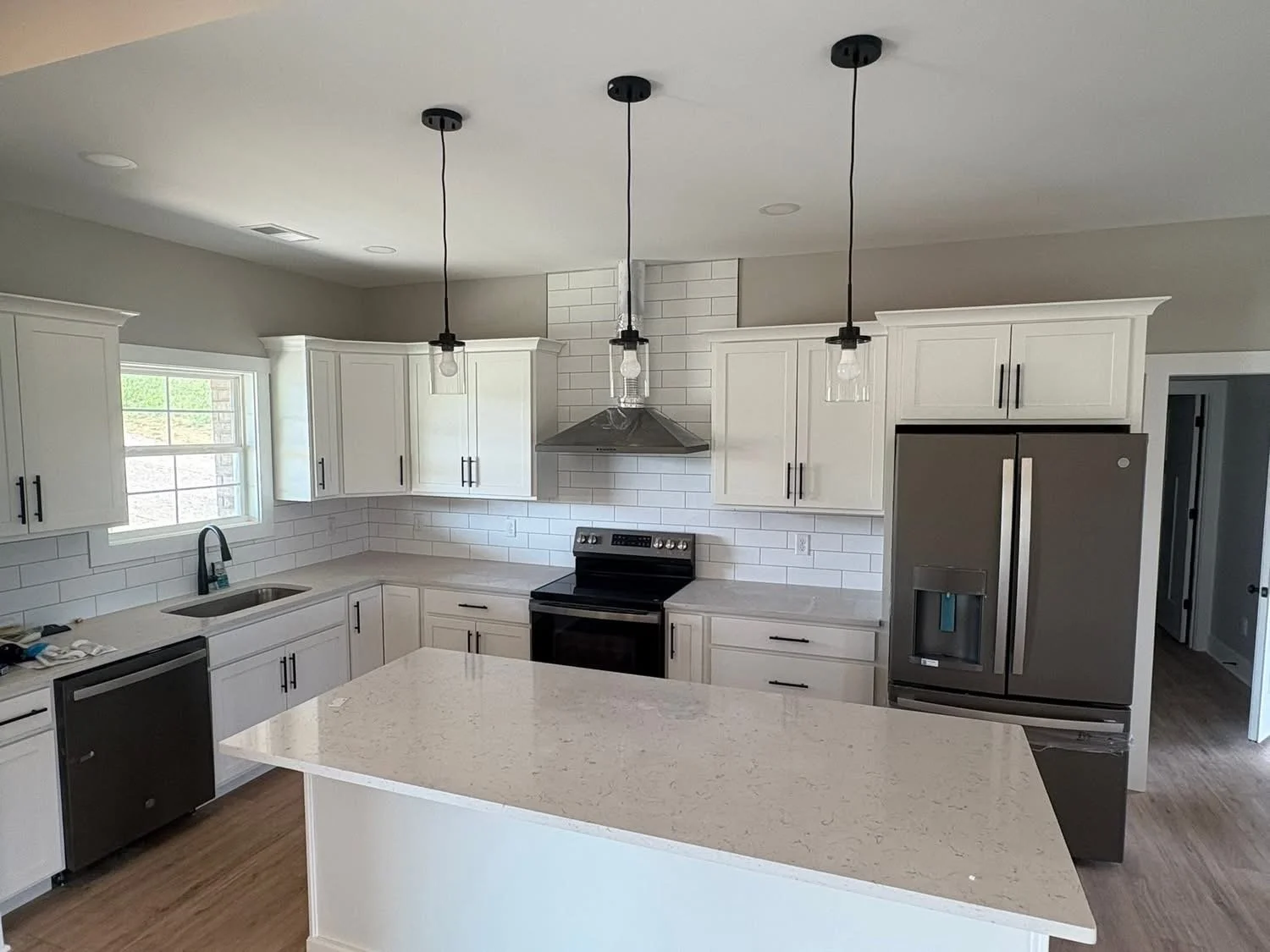 Modern kitchen with white cabinets, a large island with a light-colored countertop, stainless steel appliances including a refrigerator, oven, dishwasher, and a range hood. Pendant lights hang above the island. There is a window over the sink and recessed lighting in the ceiling.
