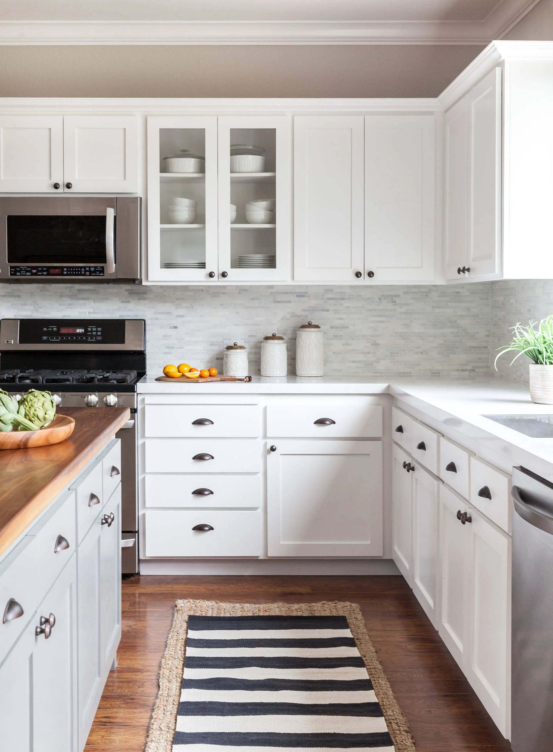 Modern white kitchen with stainless steel appliances, wooden floor, striped rug, and decorative containers, featuring upper and lower cabinets.