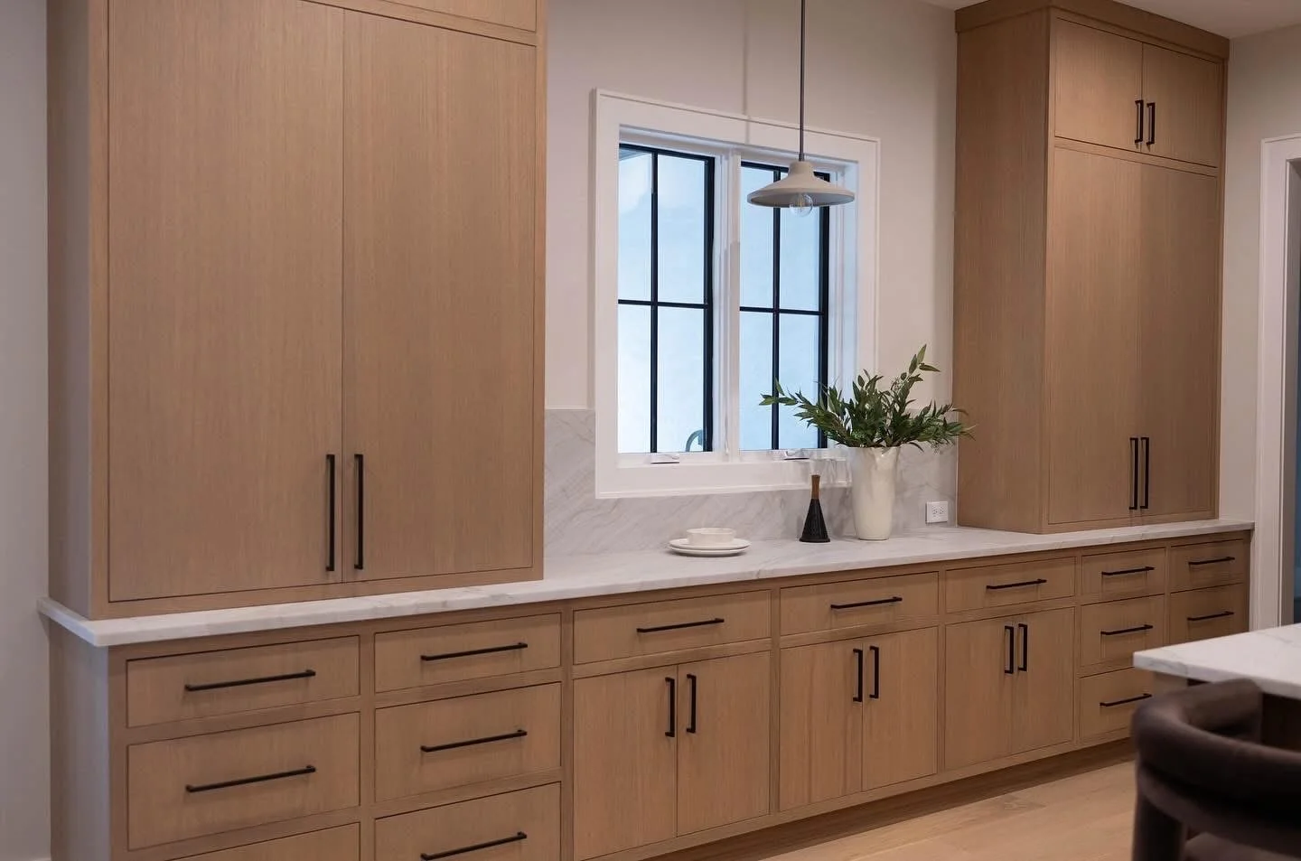 Kitchen with light wood cabinets, a marble countertop, and a window with black framing. Decor includes a white vase with green leaves, a small black vase, and stacked white dishes. There is a hanging light fixture above the window.