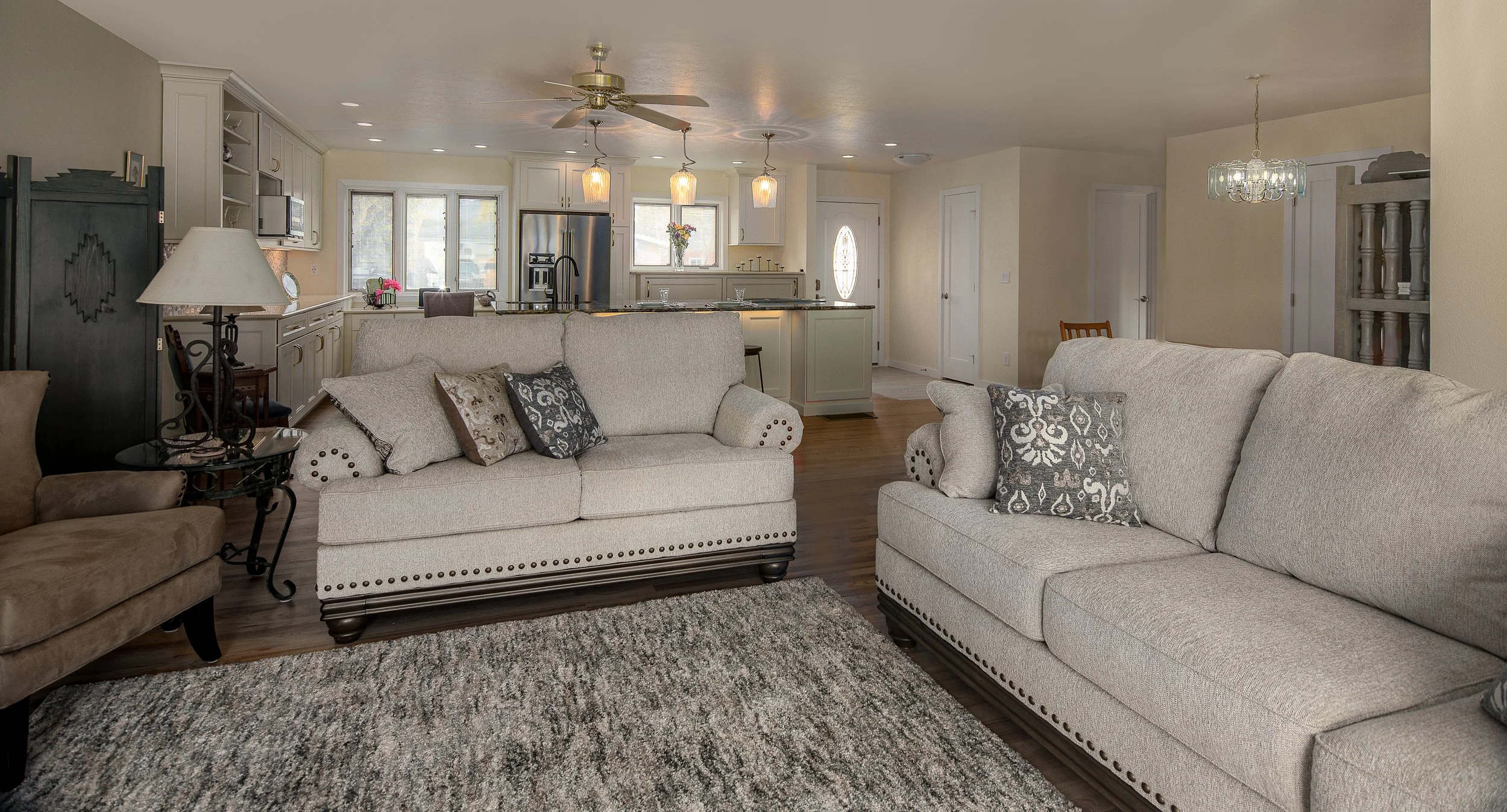 Living room with two beige sofas, a patterned rug, and a kitchen in the background featuring white cabinets, a stainless steel refrigerator, and pendant lights.
