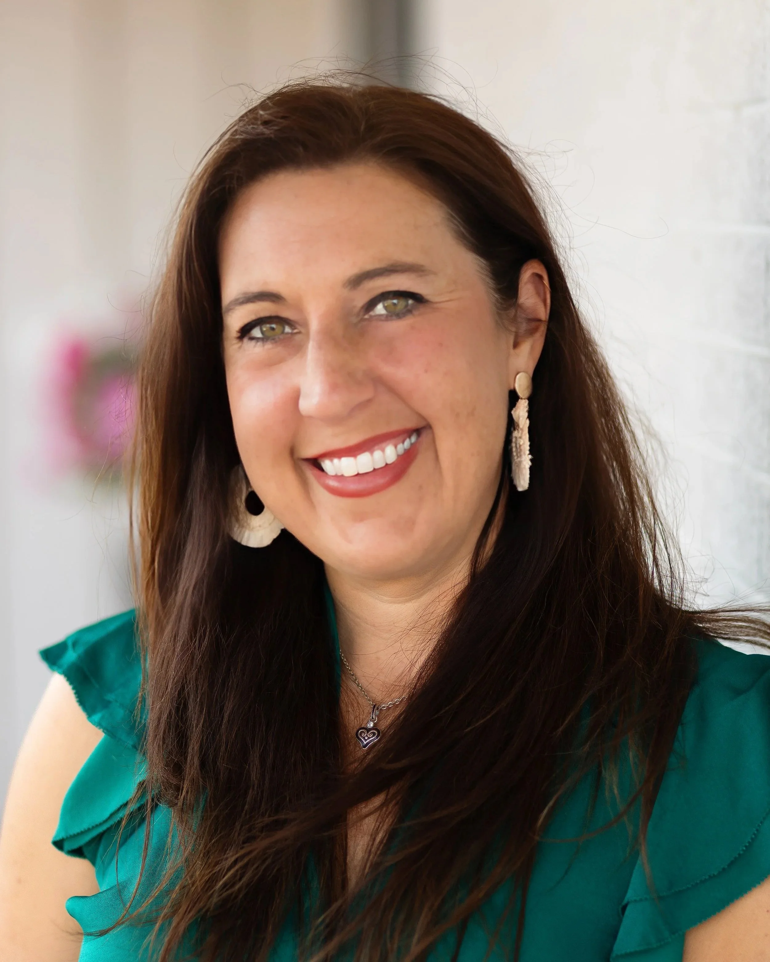 A woman with long brown hair, wearing a teal top, earrings, and a necklace, smiling in front of a blurred light background.