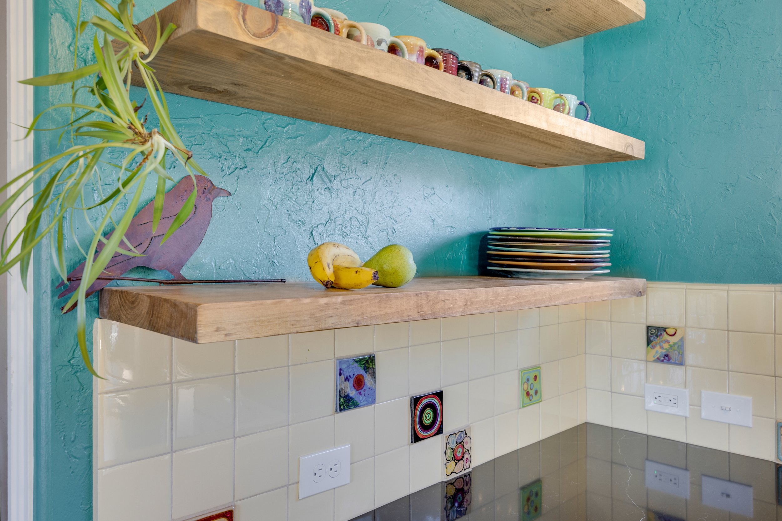 A kitchen with two wooden shelves, a turquoise textured wall, and a tiled backsplash with colorful decorative tiles. On the lower shelf, there are two bananas and a green pear, along with stacked plates. A leafy plant with purple bird-shaped decoration is on the left side.