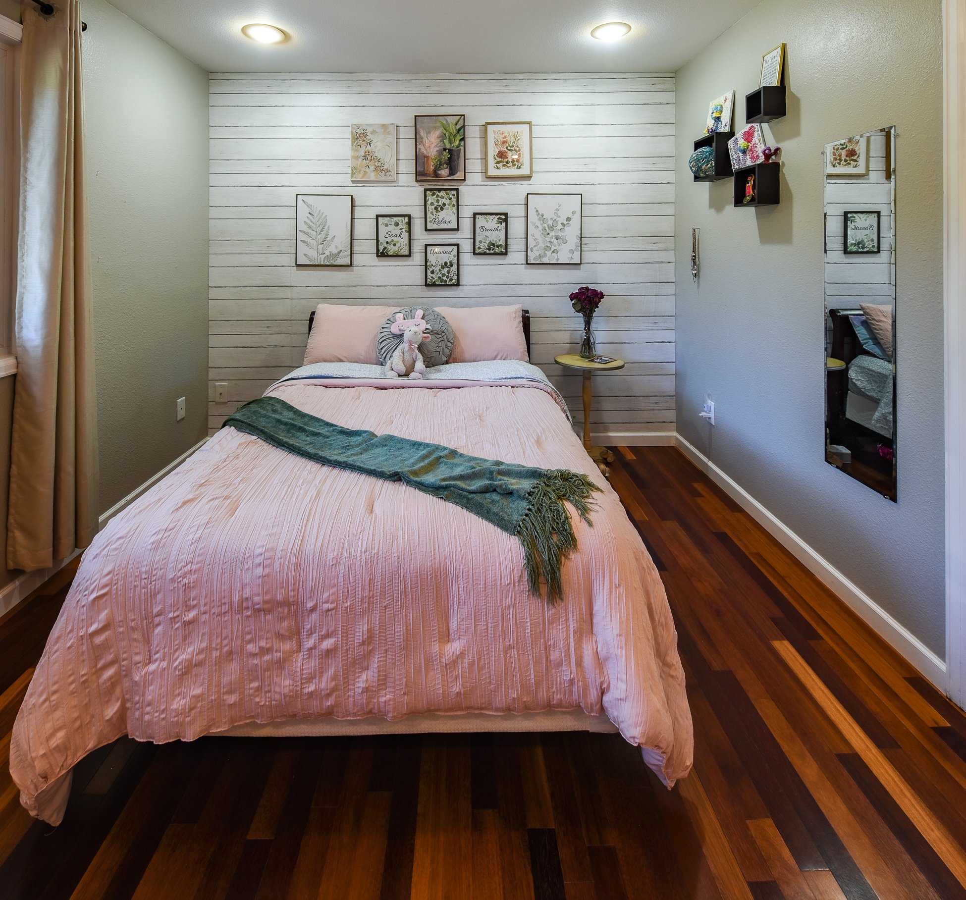 A cozy bedroom with a bed covered in pink bedding, a stuffed animal on top, and a green throw blanket. The wall behind the bed has white shiplap decor with picture frames. To the right, a small round side table with a vase of flowers and a mirror on the wall.