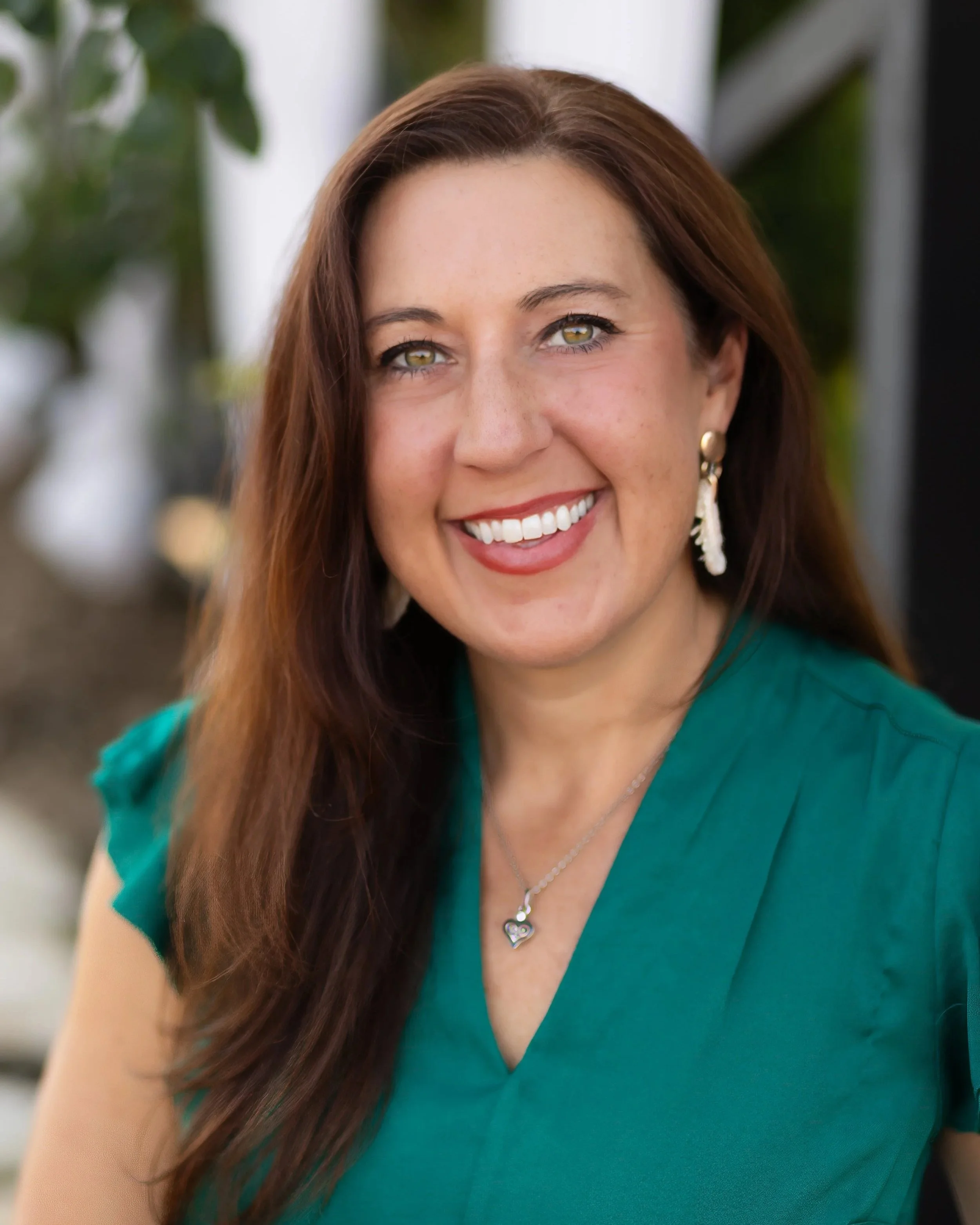 A woman with long brown hair wearing a teal top, gold earrings, and a heart-shaped pendant necklace, smiling outdoors with a blurred background.