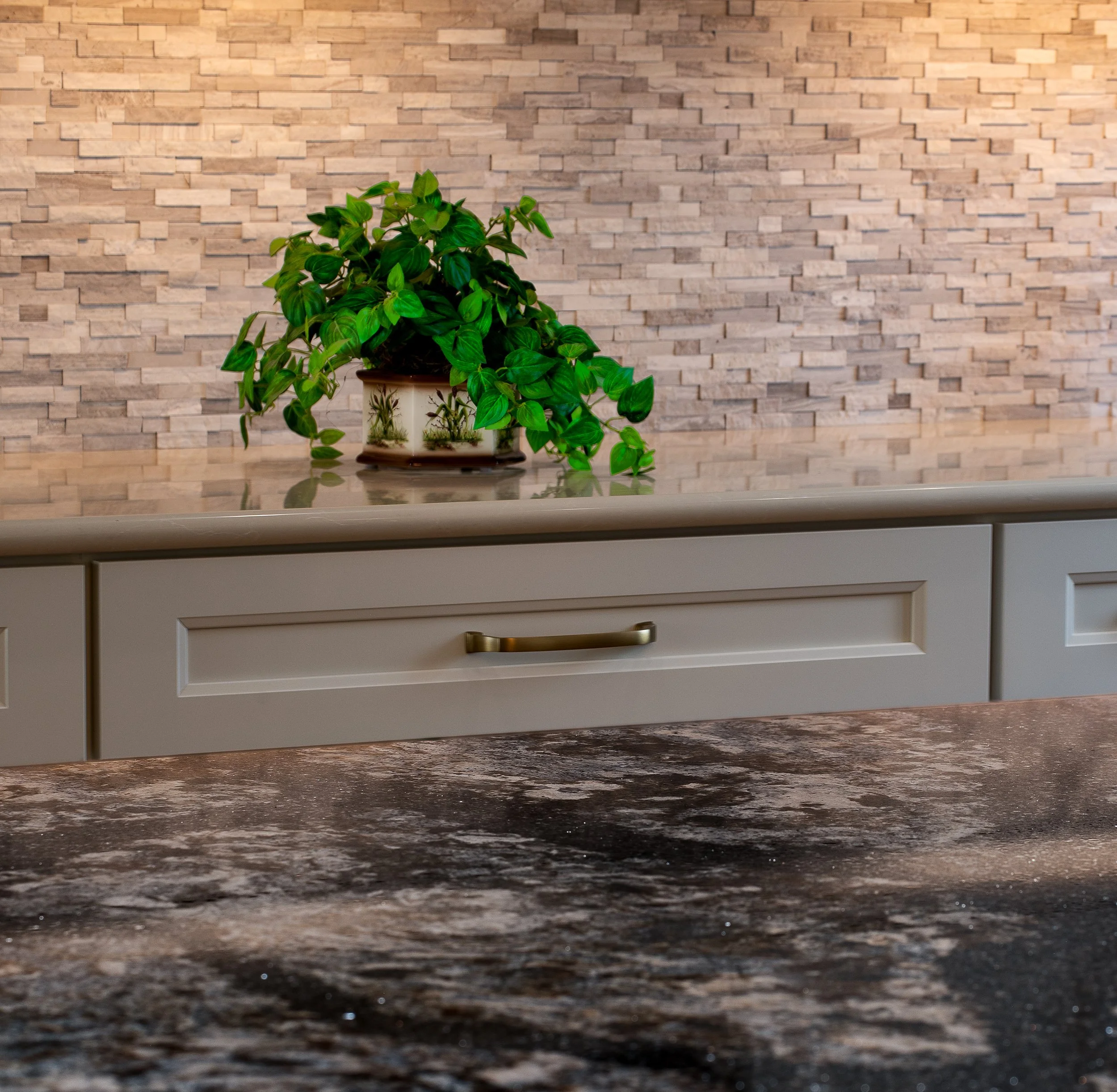 A potted green plant sitting on a kitchen counter with a beige drawer and a brick wall in the background.