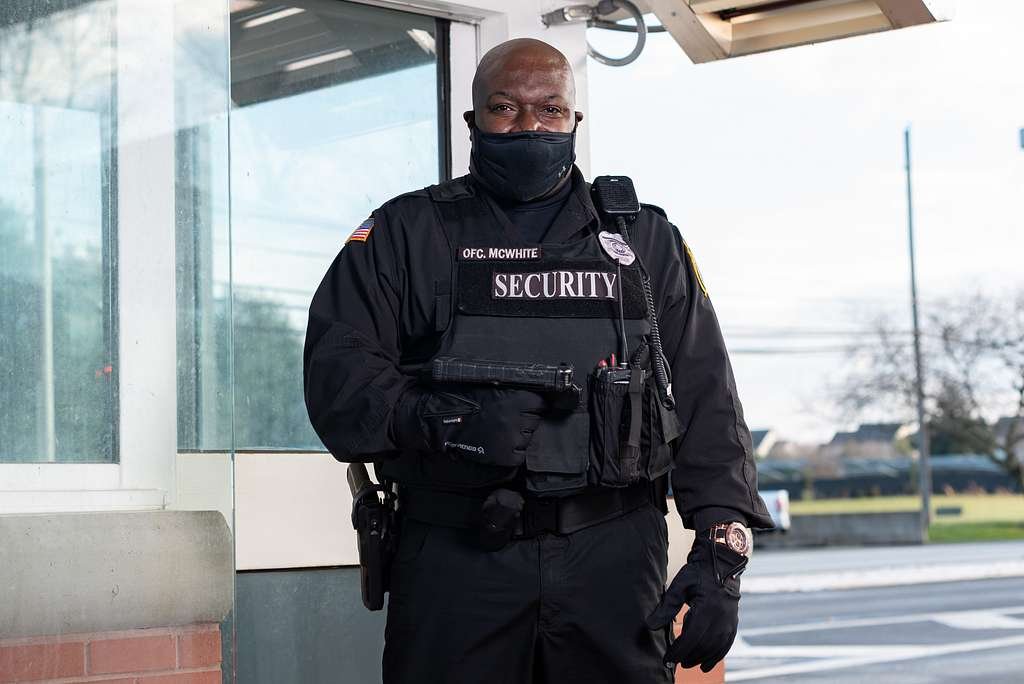 A security officer standing outdoors in front of a building, wearing black tactical gear, a black mask, and a watch.