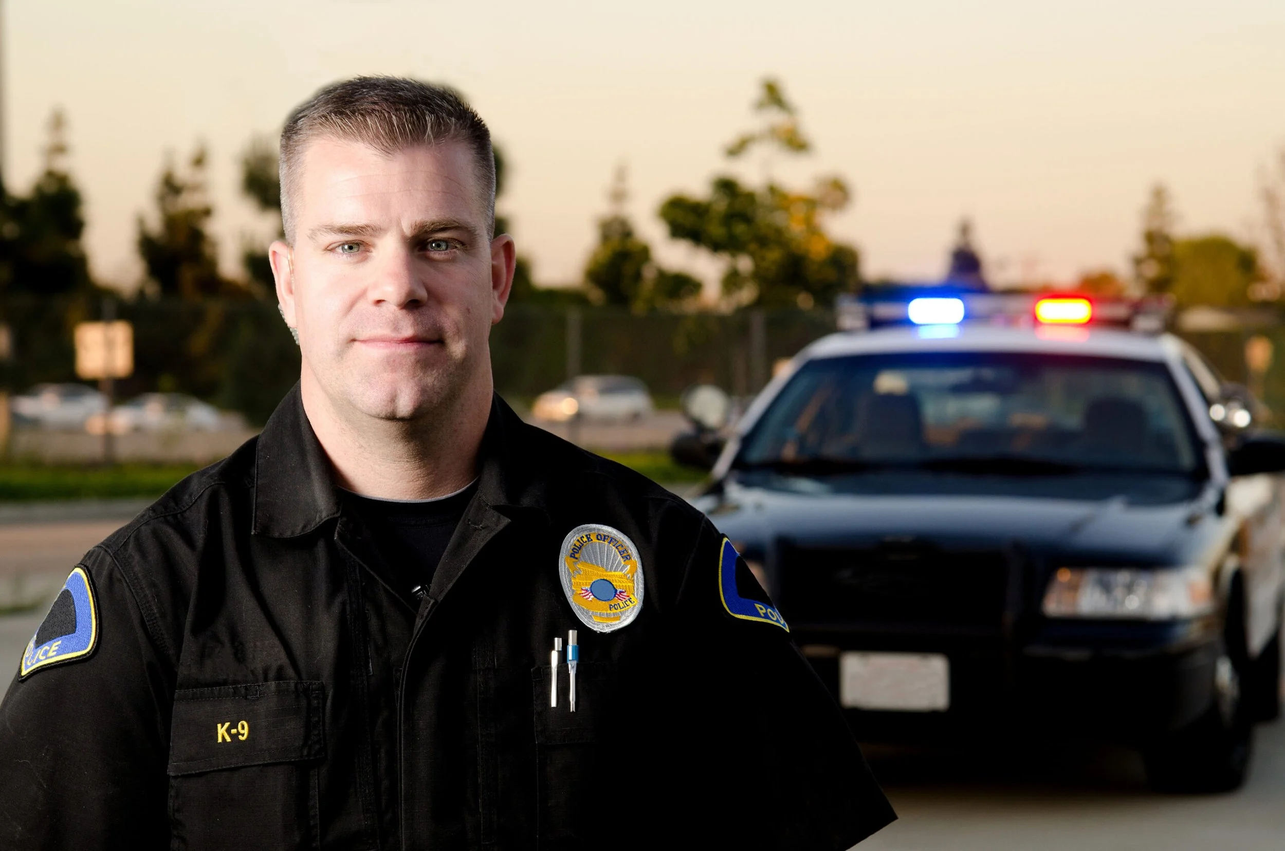 A police officer standing in front of a police car with flashing lights during sunset.
