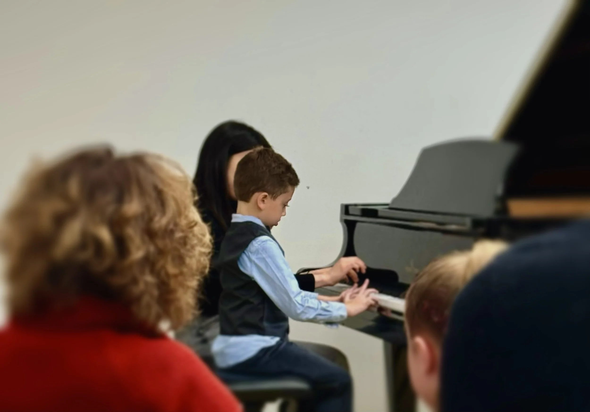 A young boy playing the piano in front of an audience while a woman, possibly a piano instructor, assists him. Several people are watching, with the focus on the boy and the woman.