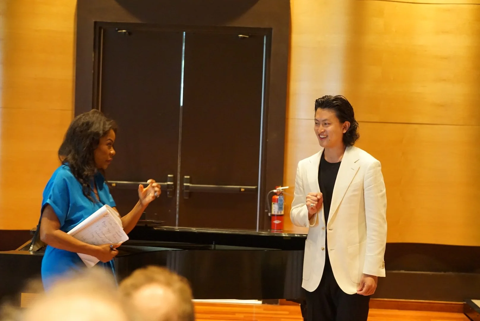 Two people having a conversation near a black grand piano in a room with warm wooden paneling. One person is a woman holding a notebook and pen, wearing a blue dress, and the other is a man wearing a white blazer and black shirt, smiling.