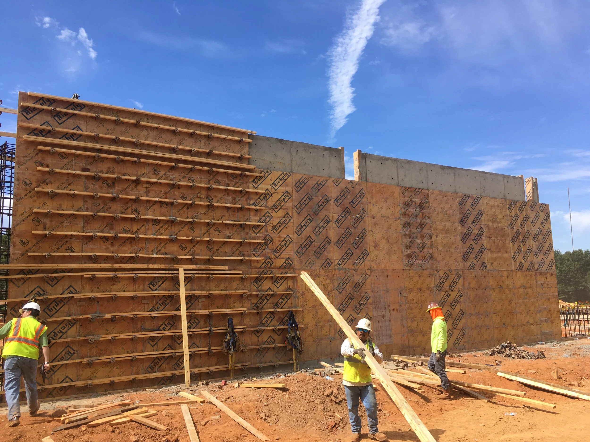 Construction workers in safety gear building a concrete wall with wooden formwork at a construction site under a blue sky.