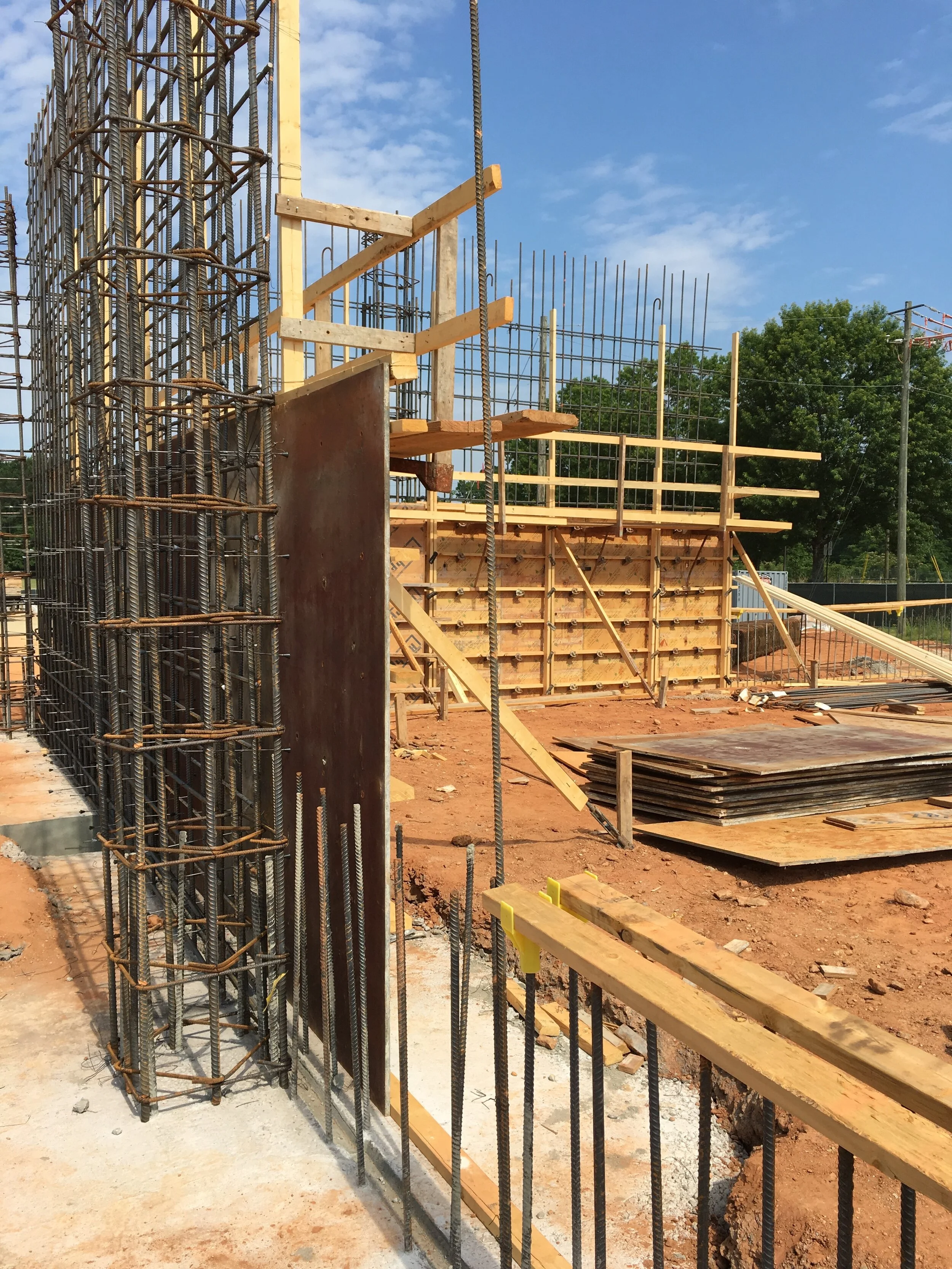 Construction site with steel rebar, wooden formwork, and scaffolding on reddish dirt ground under a blue sky with some clouds.