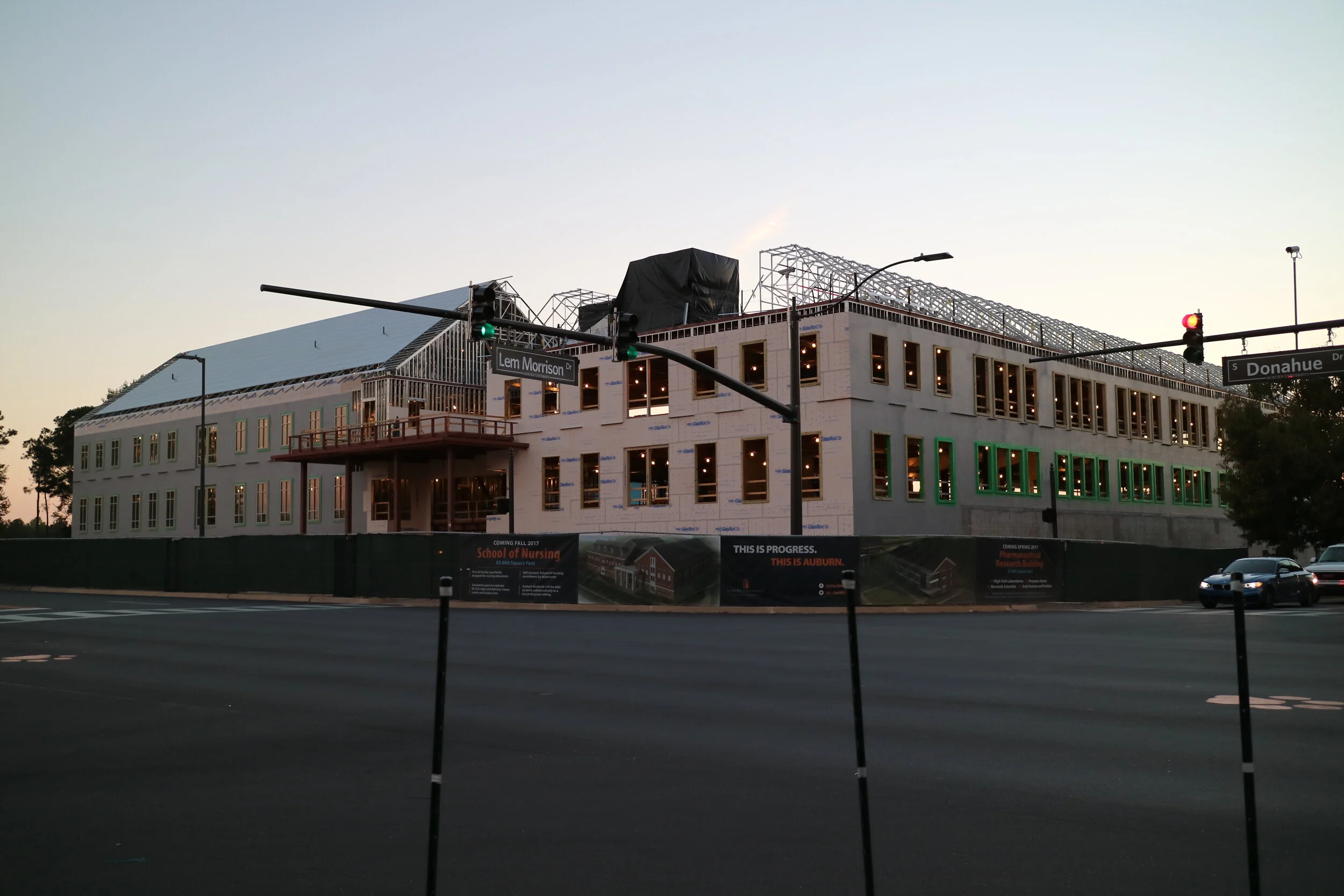Under construction building at the corner of Lem Morrison Drive and Donahue Drive, with signs indicating it is a School of Nursing in Auburn, surrounded by streetlights and cars during dusk.