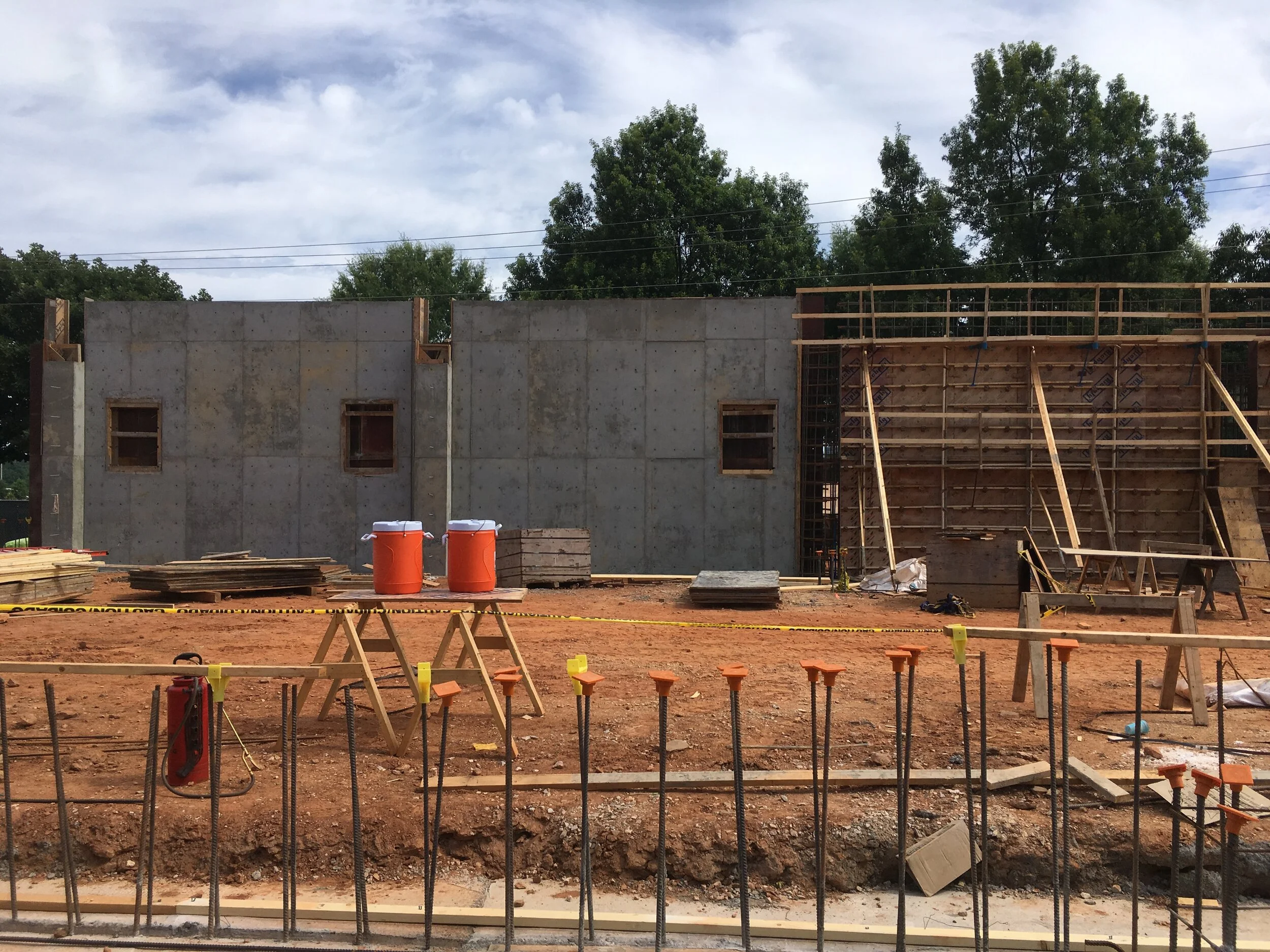 Under construction building with concrete walls, wooden scaffolding, orange safety barrels, and construction tools on dirt ground.