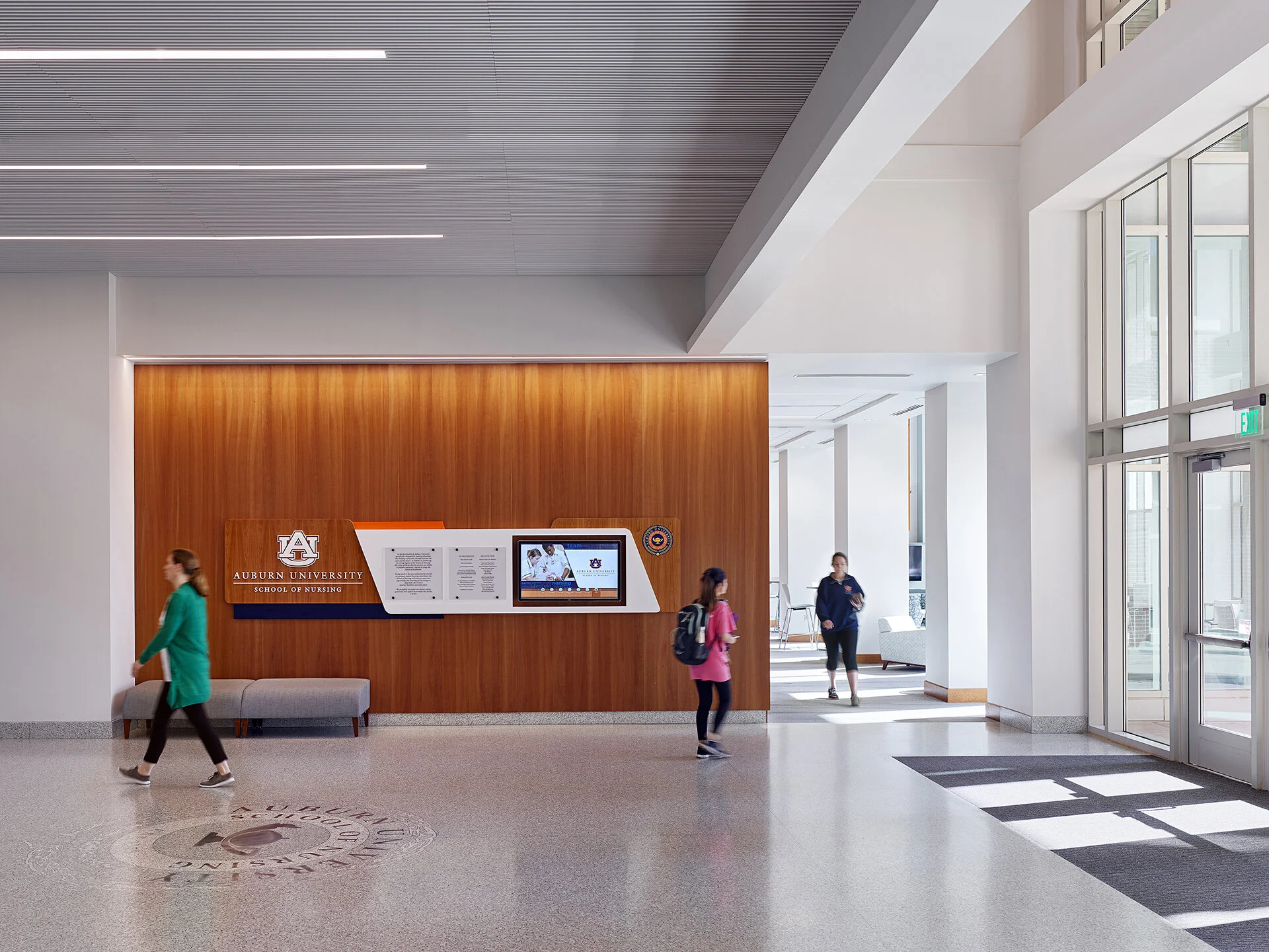 Interior view of Auburn University School of Nursing reception area with people walking by, a wooden wall with Auburn University logo and information display, large windows, and a glass door.