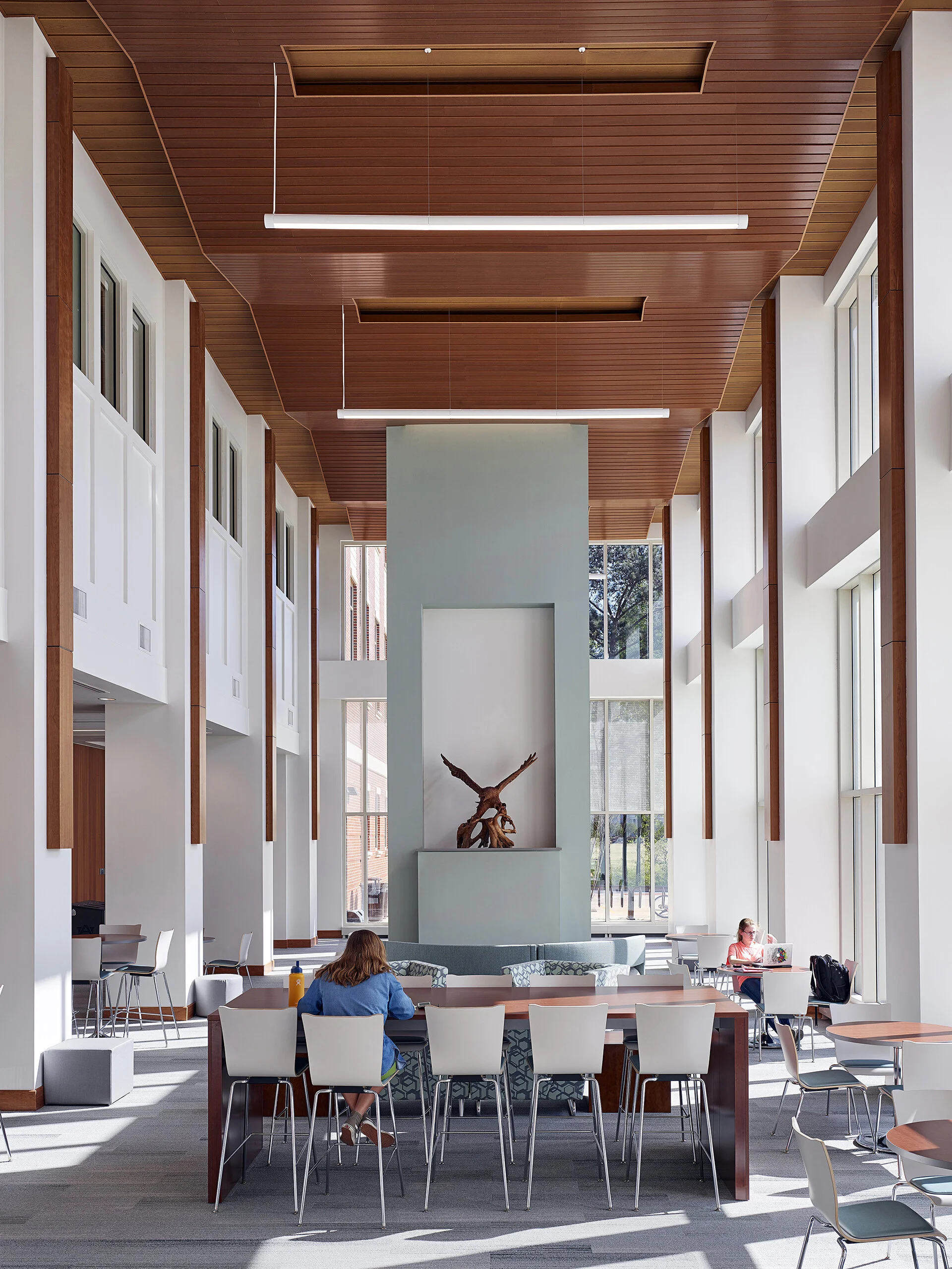 Interior of a modern public space with high ceilings, large windows, wooden accents, and a central sculpture of an eagle with wings spread, surrounded by white chairs and tables.