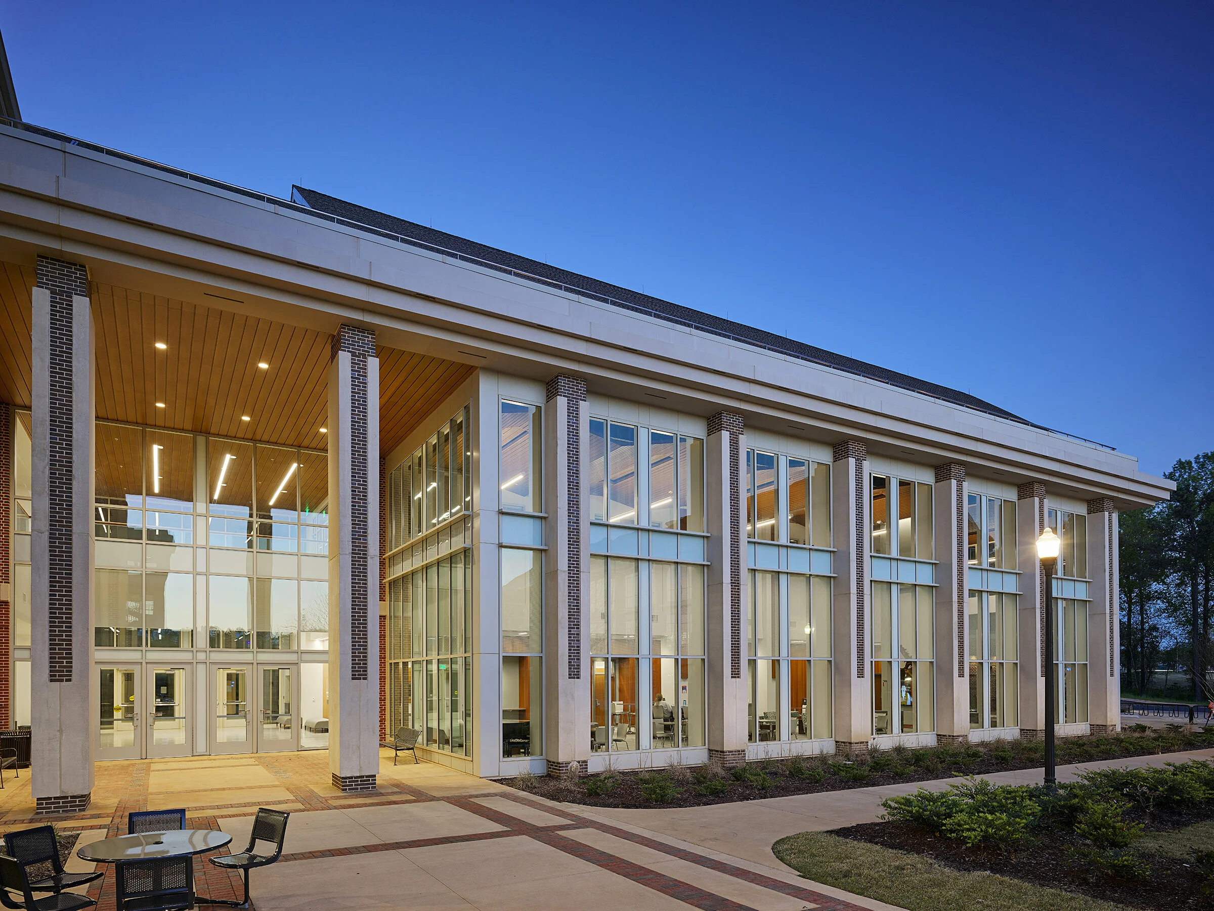 Modern two-story building with large glass windows, brick columns, and a wooden ceiling over the entrance, exterior garden, and outdoor seating area, taken during early evening.