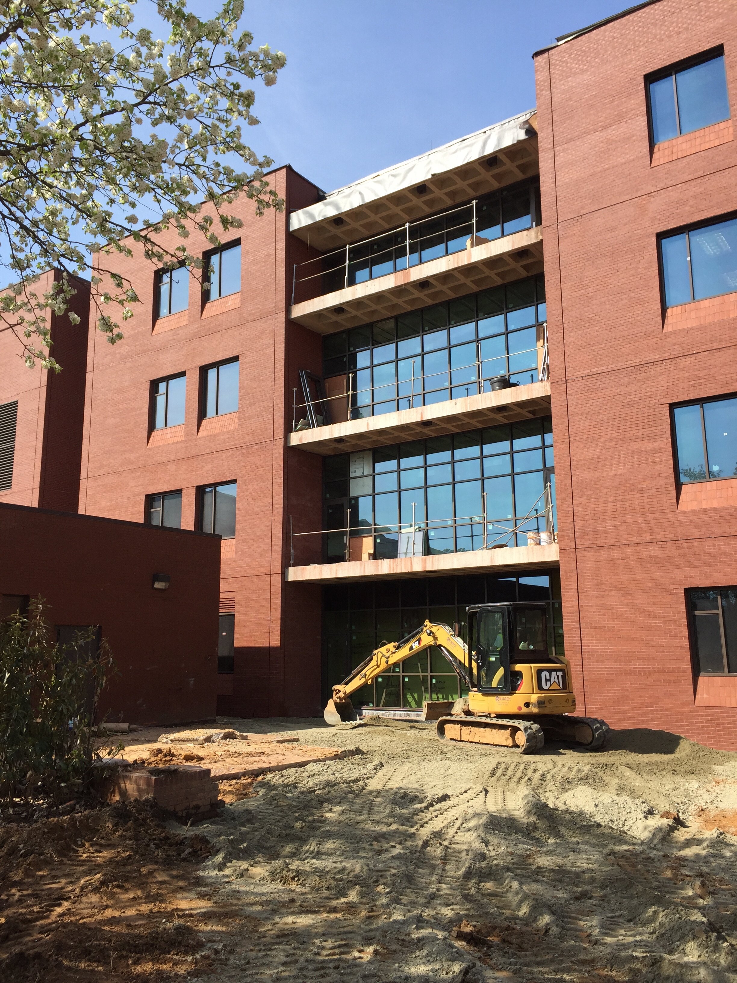 Construction site with a small yellow excavator in front of a multi-story brick building that has large glass windows and balconies under construction.