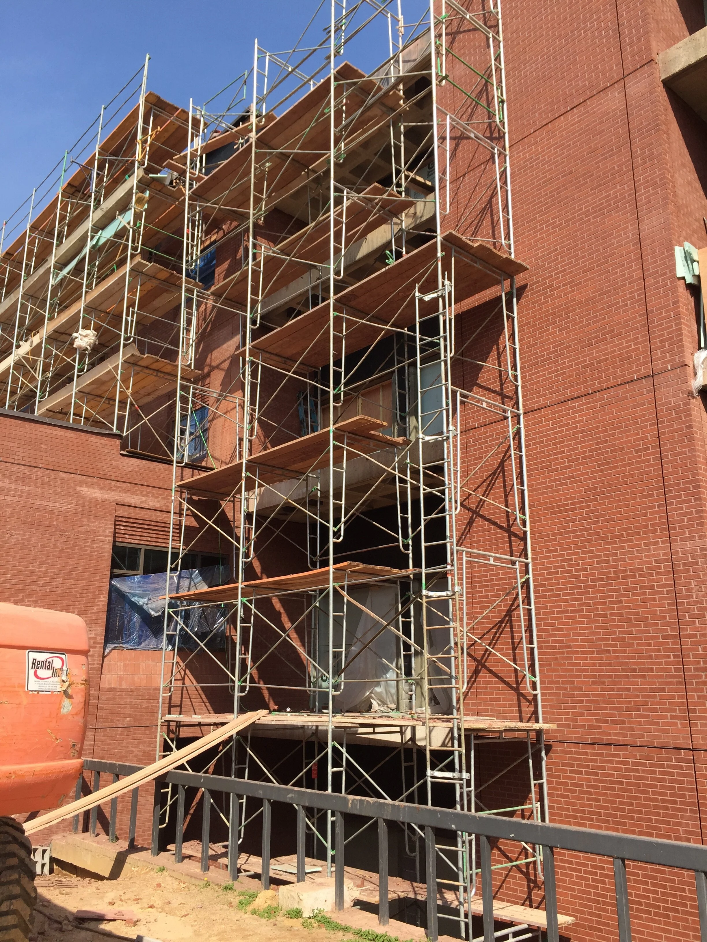 Construction scaffolding set up on a brick building under renovation, with some windows covered in plastic. A construction lift is visible in the foreground.