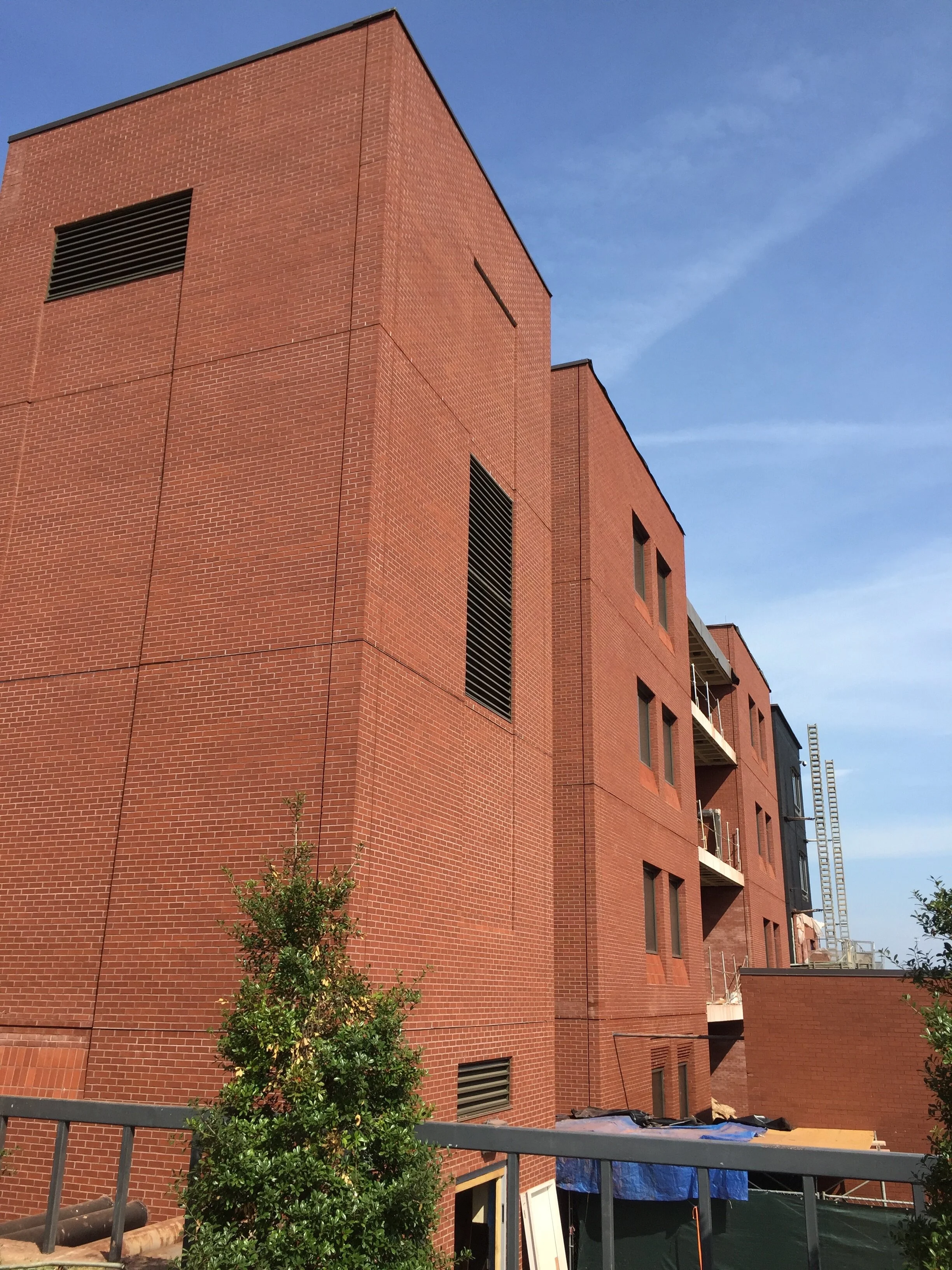Red brick building under construction with windows and balconies, blue sky, green trees, and construction materials in the foreground.