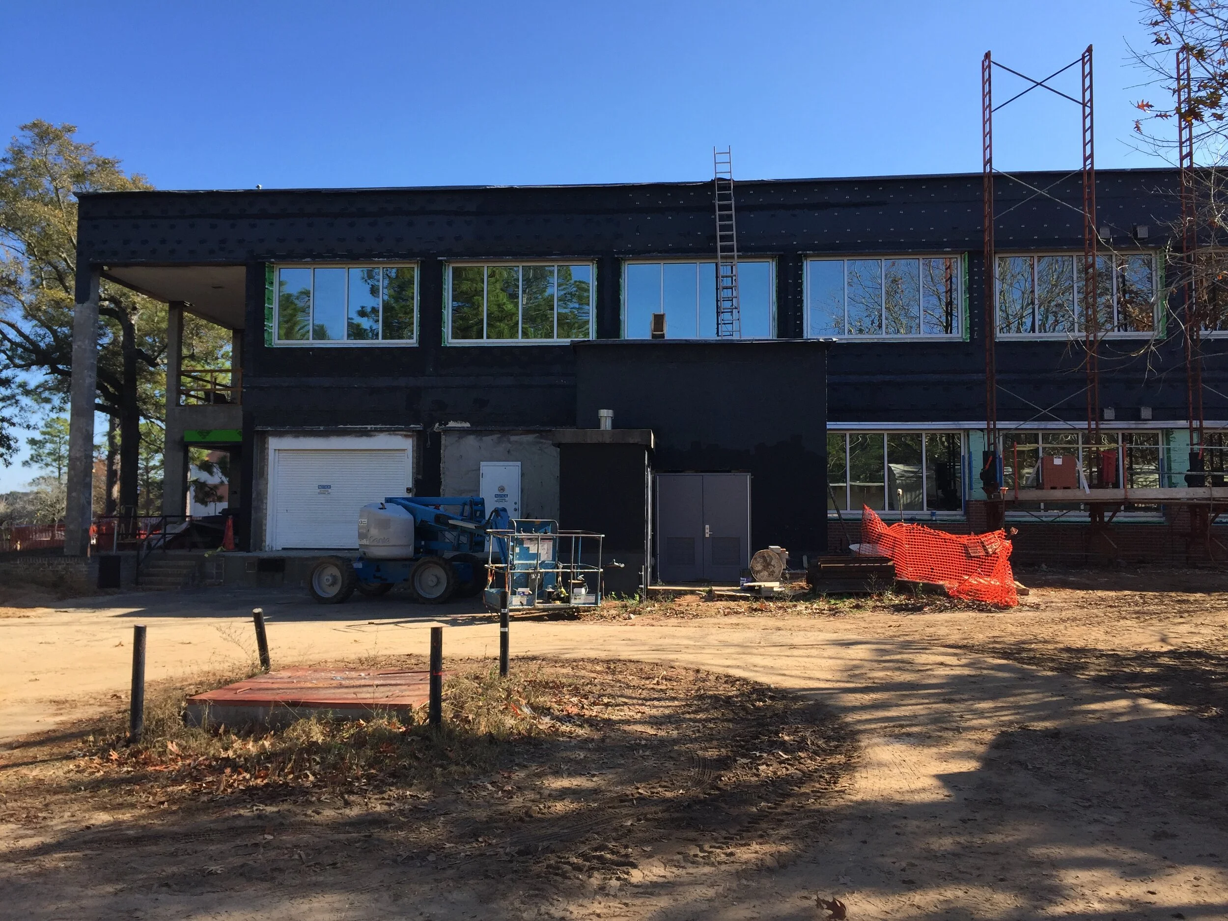 Construction site with a black building under construction, scaffolding, and equipment. The building has large windows, and the ground is dirt with some construction barriers.