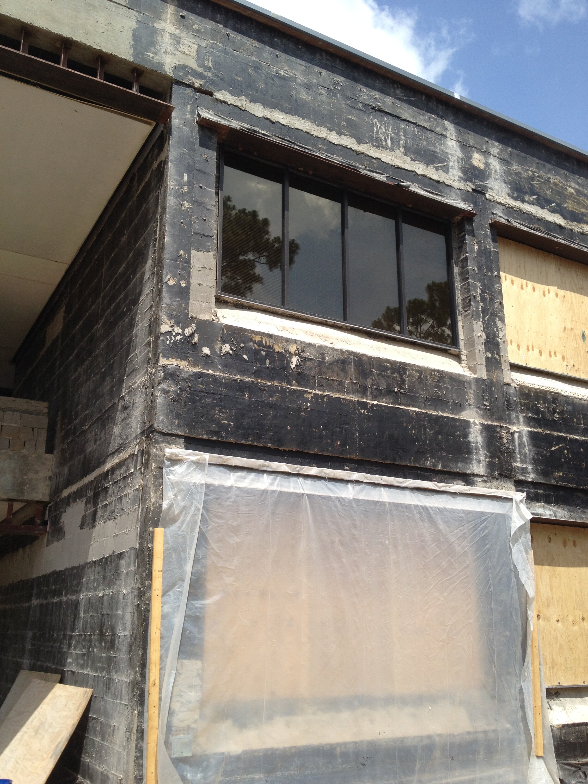 Construction site of a building with black brick exterior, a large window with black frame, and temporary plywood covering a carport or garage area.