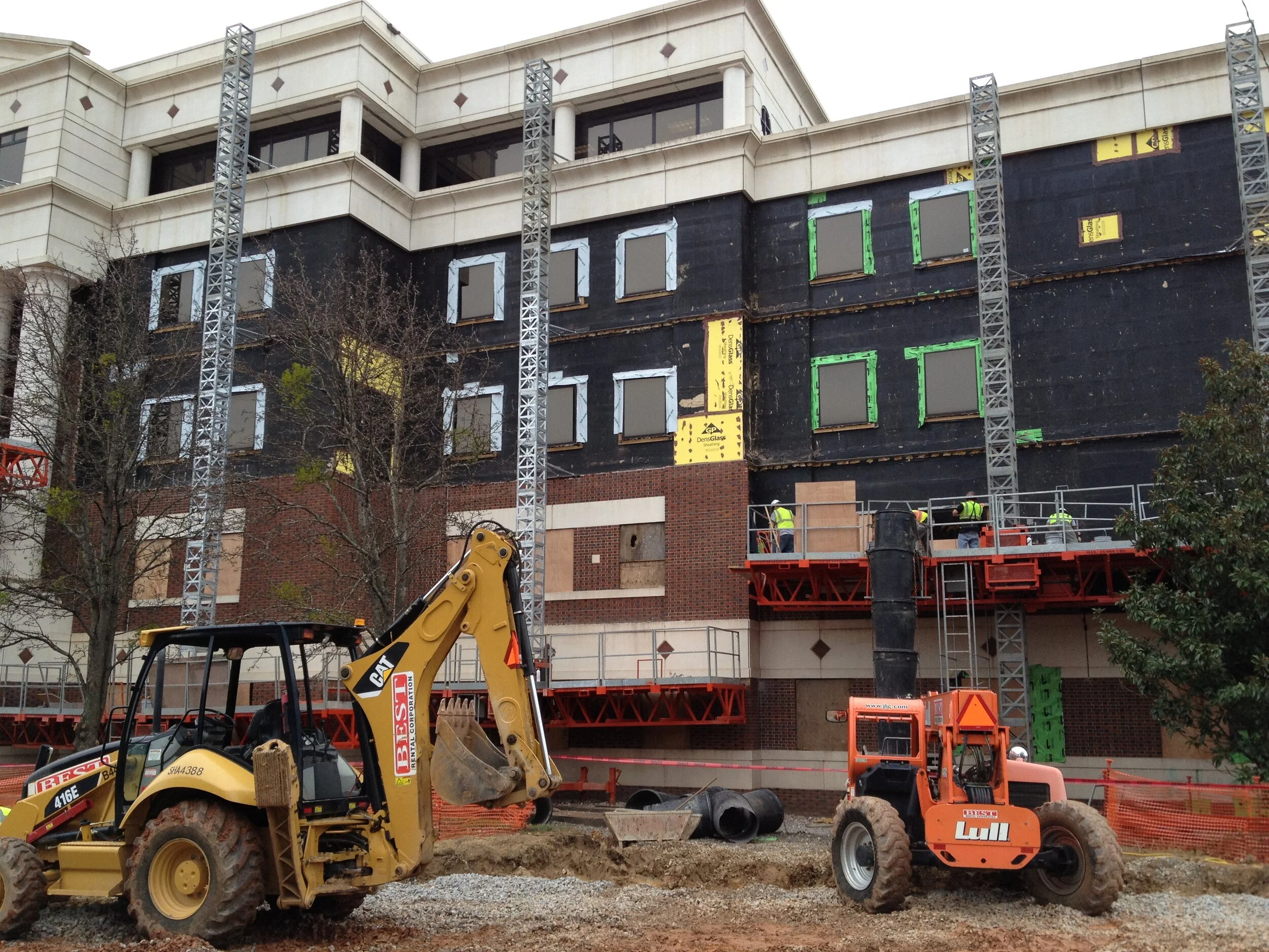 Construction site with workers installing or repairing windows on a multi-story building. Heavy machinery such as a backhoe and a roller are present in the foreground.
