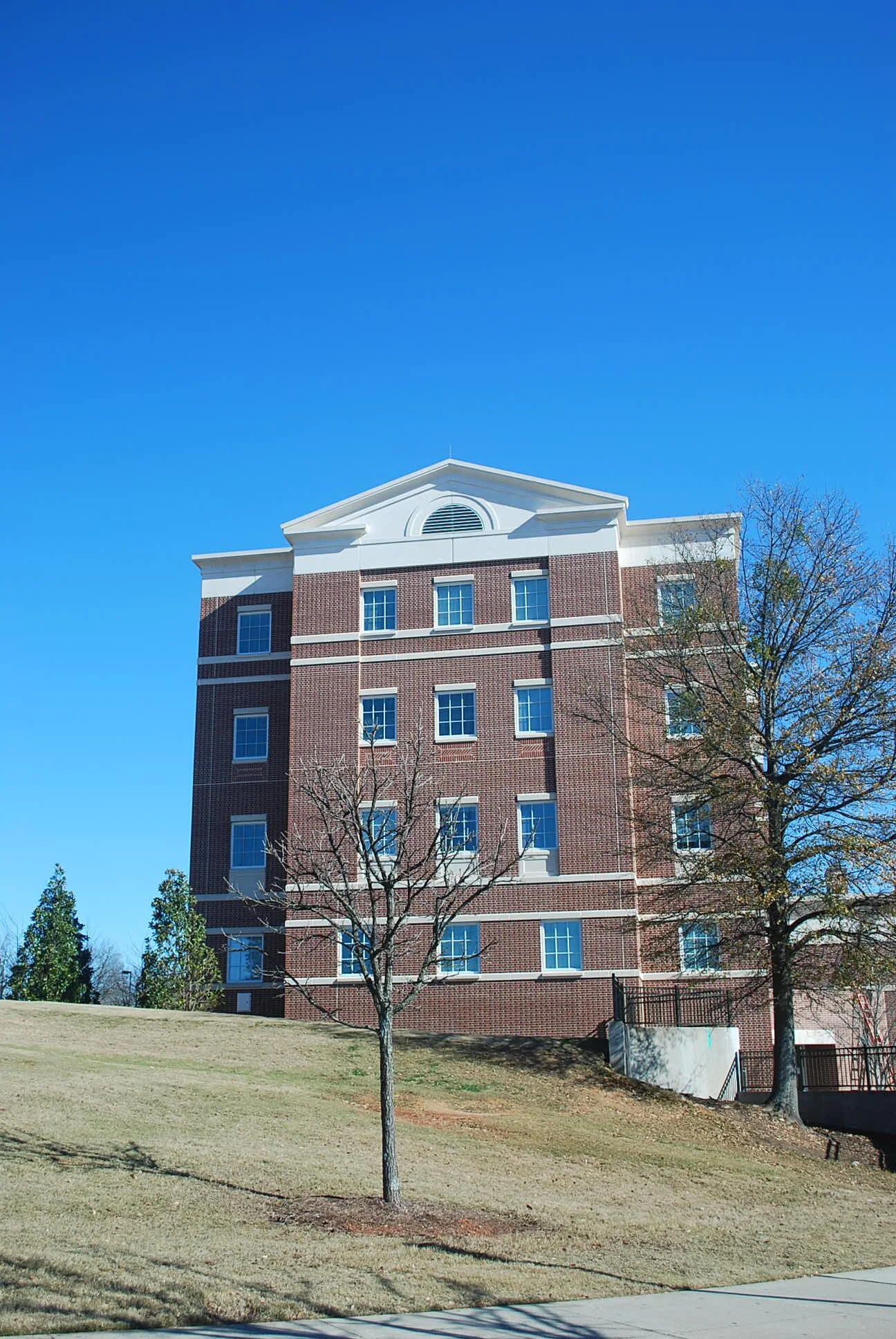 A multi-story brick building with white trim under a bright blue sky, with leafless trees and a grassy area in the foreground.