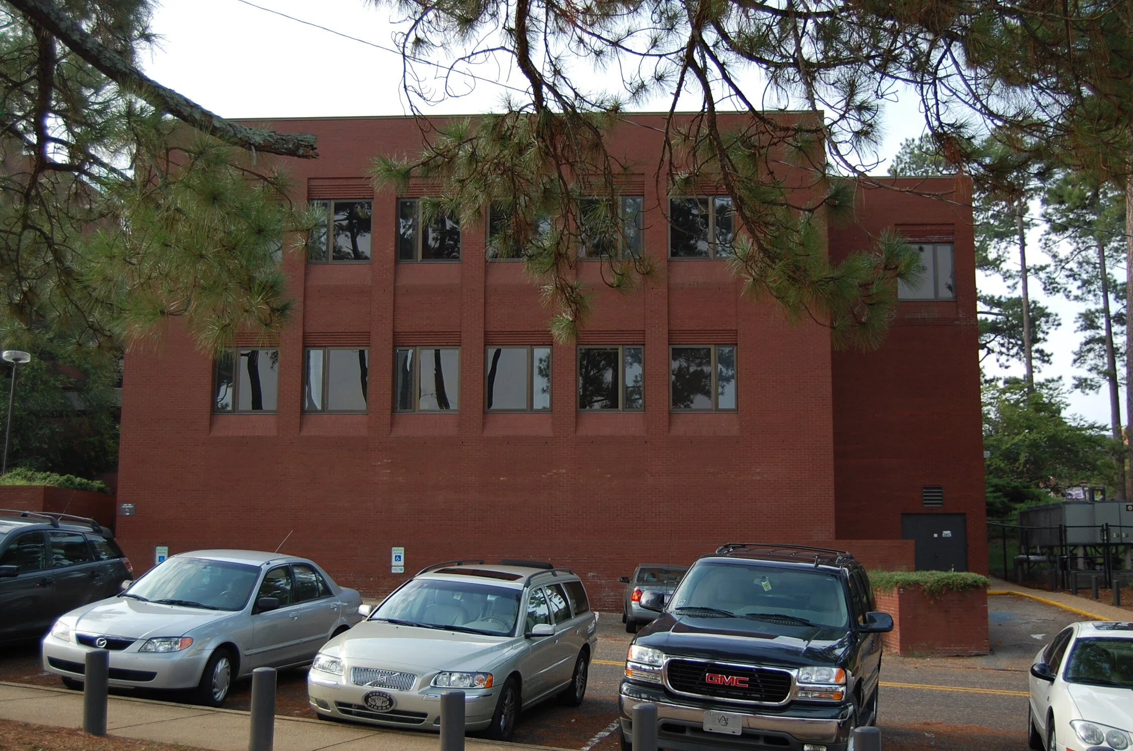 A brick building in a parking lot with several cars parked in front, trees partially obscuring the view.