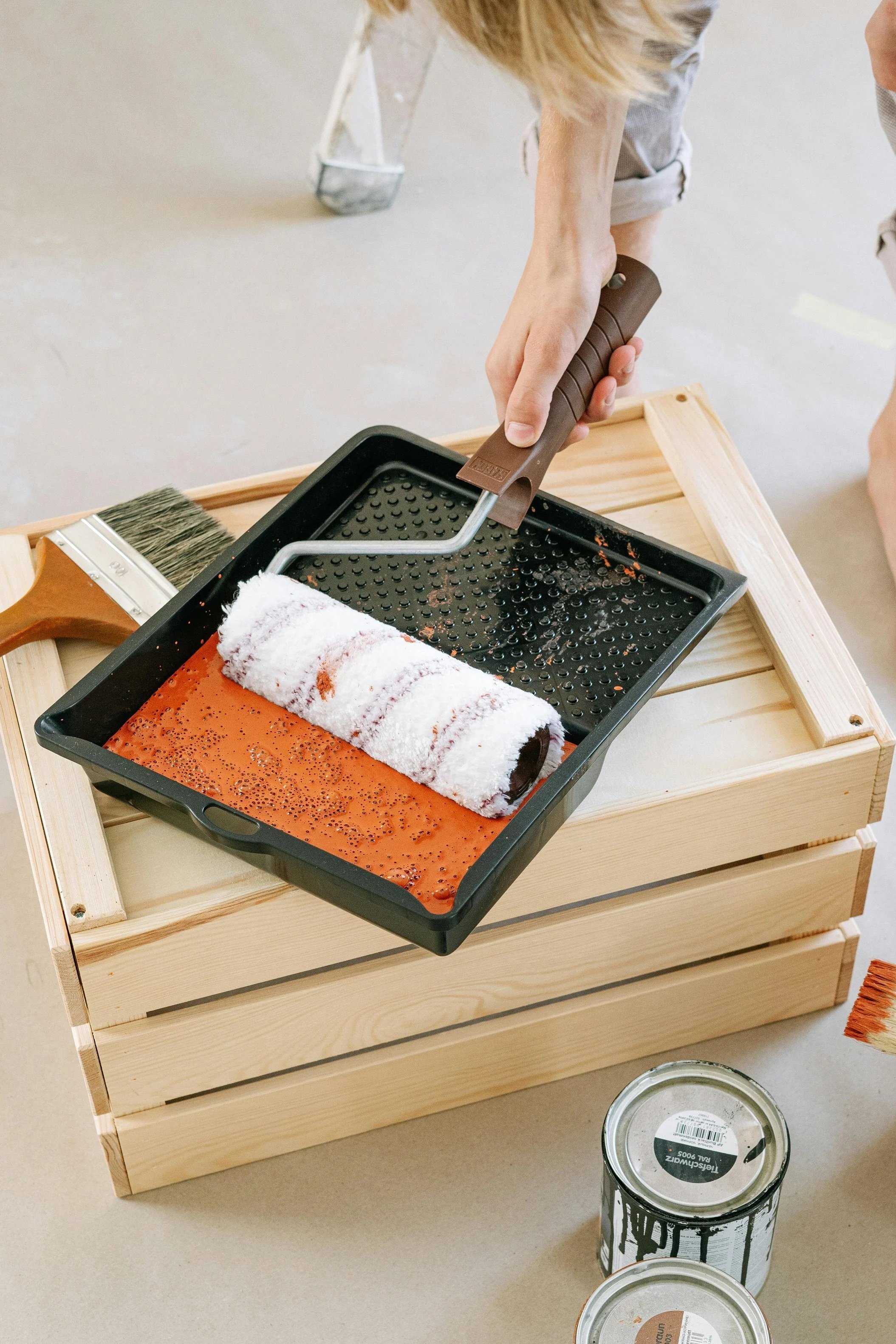 A paint roller in a tray with Benjamin Moore orange paint with and a paintbrush on a box