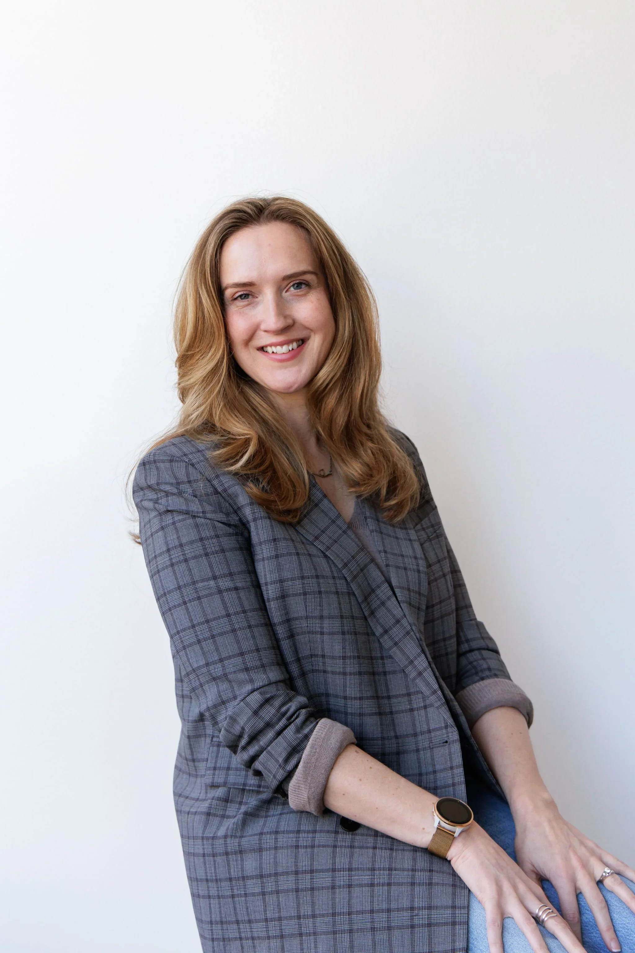 A woman with long wavy red hair smiling, wearing a gray plaid blazer and a smartwatch, standing against a plain white wall.