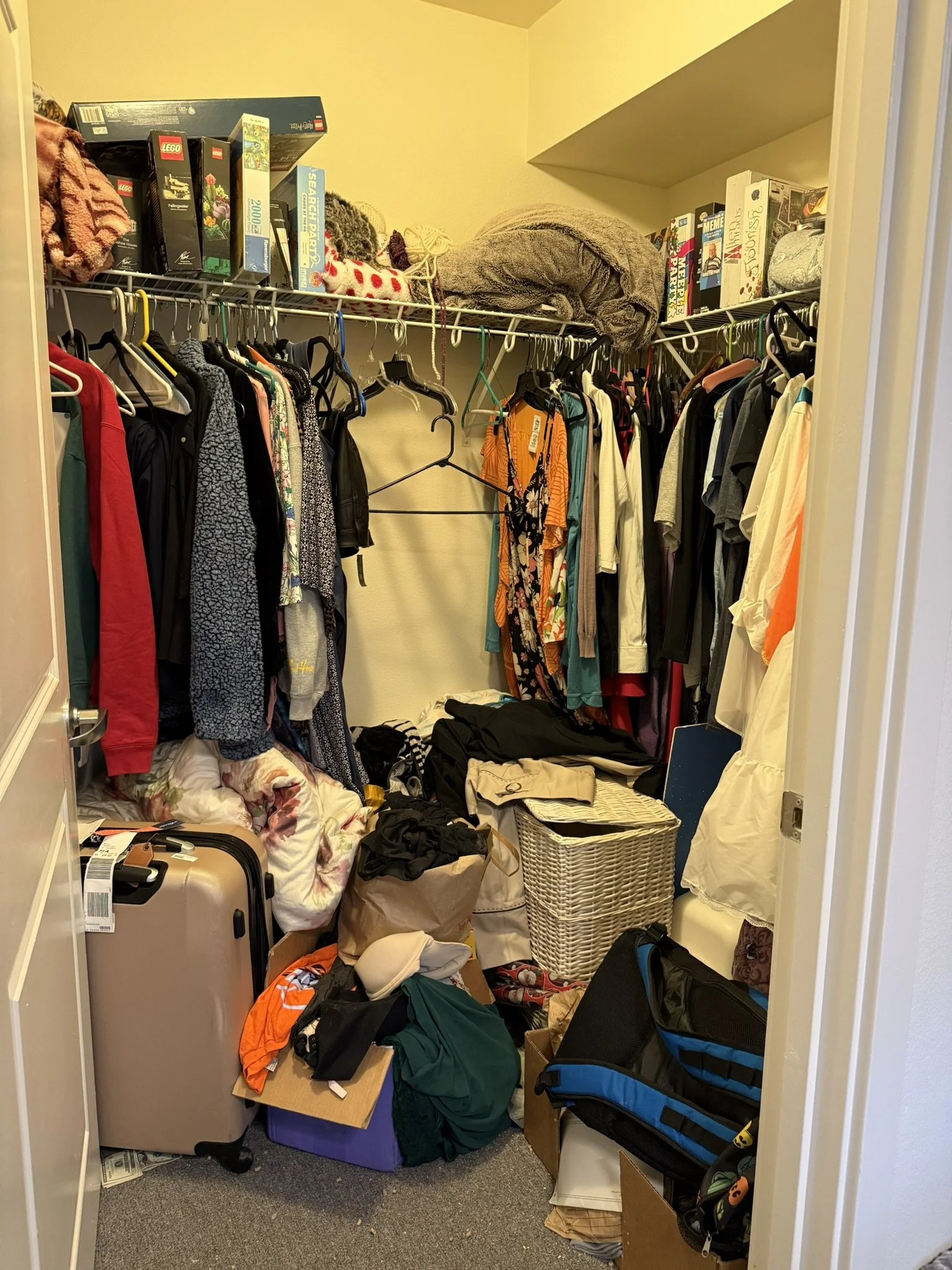 A cluttered walk-in closet filled with hanging clothes, boxes, a suitcase, and various household items.