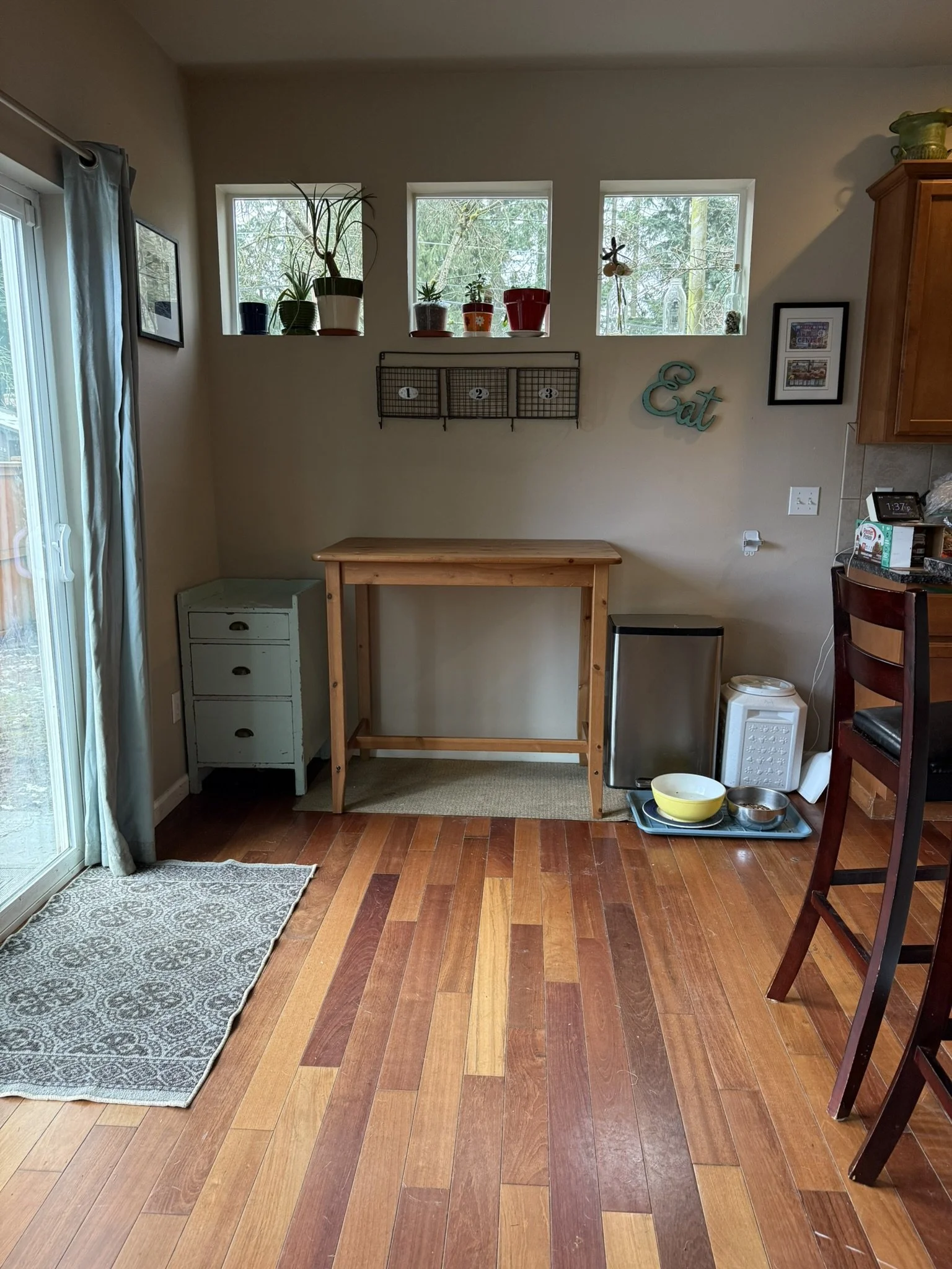 A kitchen nook with three small windows above, decorated with potted plants and a wall-mounted wire rack. There is a wooden table, a gray small cabinet, and a tray with bowls and pet food containers on the floor. Part of a sliding glass door with cur