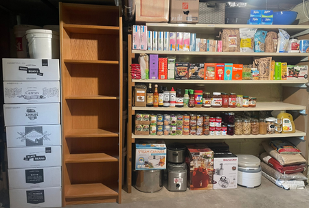 Empty wooden pantry shelf next to filled pantry shelves with food and supplies including canned goods, boxes, jars, and household items