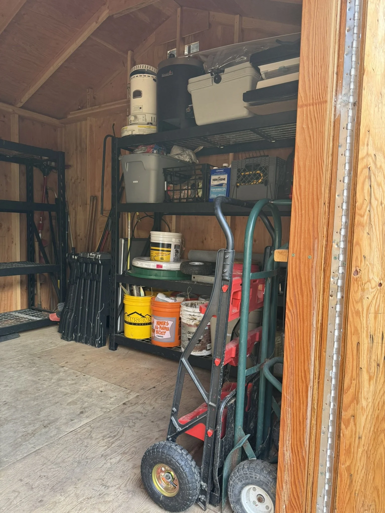 A storage shed with metal shelves filled with various containers, tools, and supplies. There are plastic buckets, a hand truck, and shelving units with boxes, cans, and other items.