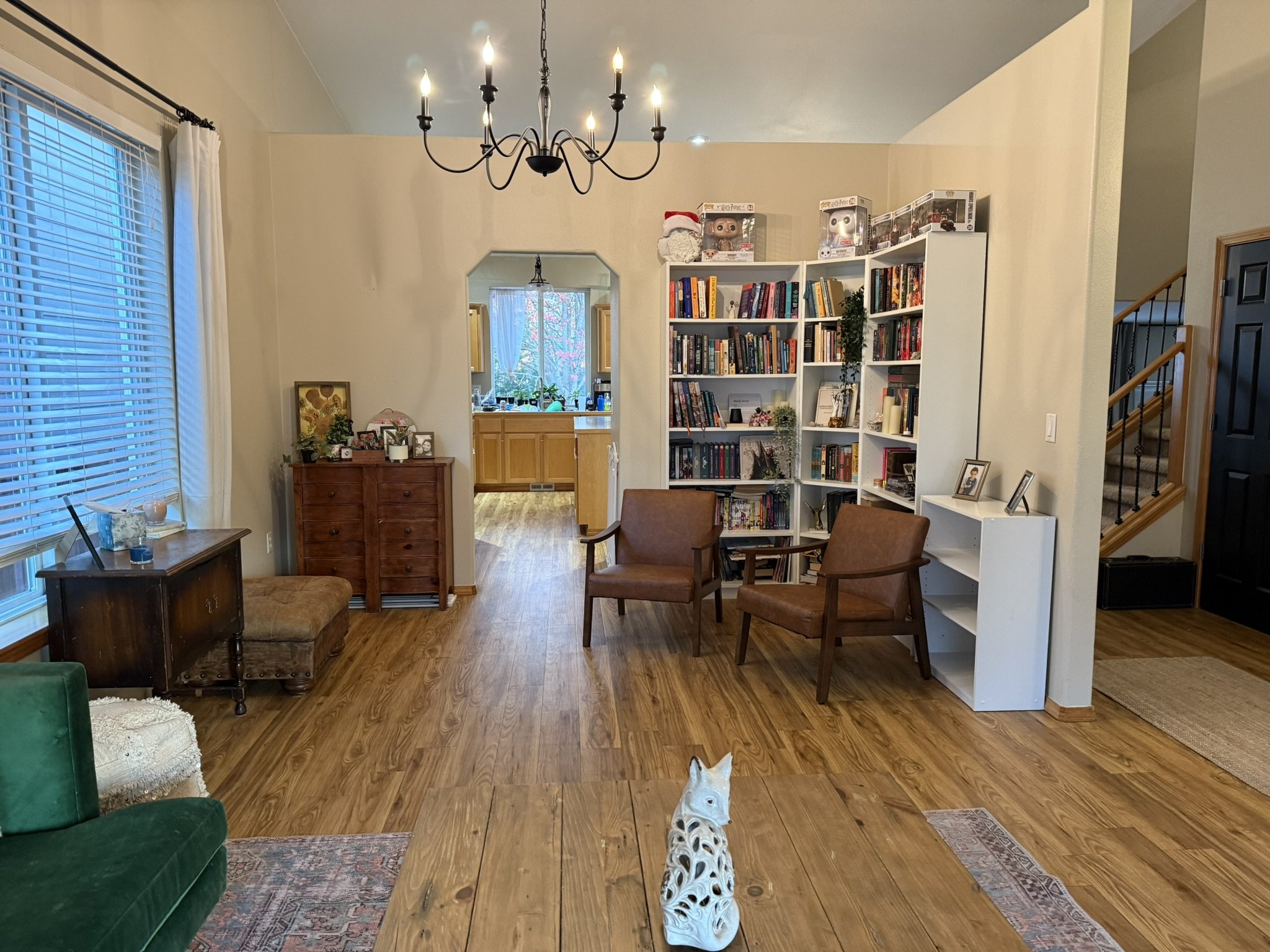 Living room with hardwood floors, a green armchair, a wooden table with a laptop and a candle, two chairs, a bookcase filled with books, and a chandelier hanging from the ceiling. There's a window with blinds on the left and a staircase on the right.