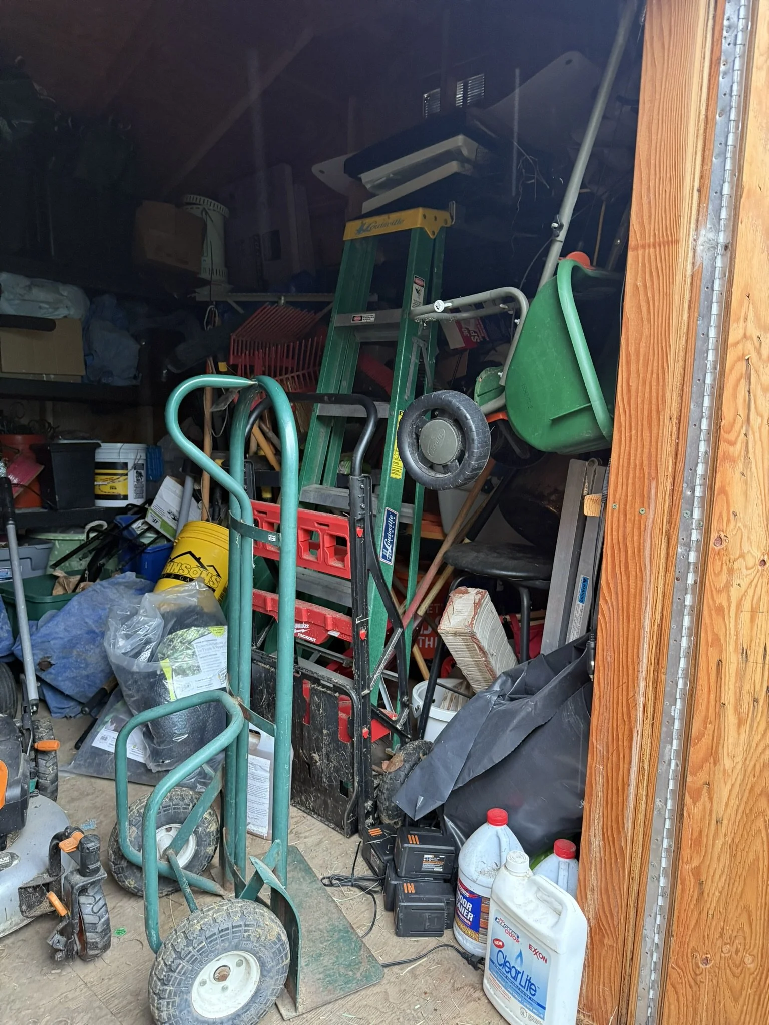A cluttered garage filled with various tools, supplies, and yard equipment, including a green wheelbarrow, a hand truck, a ladder, and containers of motor oil, cleaning supplies, and other items.