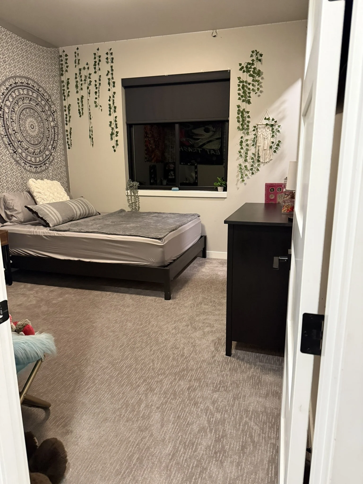 A bedroom with a bed on the left, decorated wall art, and hanging artificial vines on the walls. There is a window with a black roller shade, and a black dresser on the right with some decorations on top. The room has beige carpet and neutral-colored