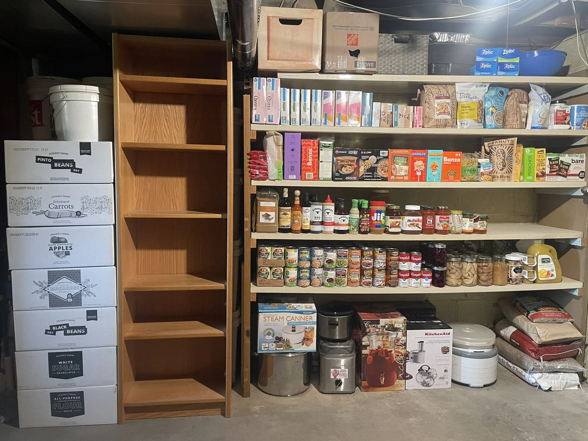 Shelves and boxes in a basement storage room with organized food and household items, including canned goods, snacks, baking supplies, and kitchen appliances.