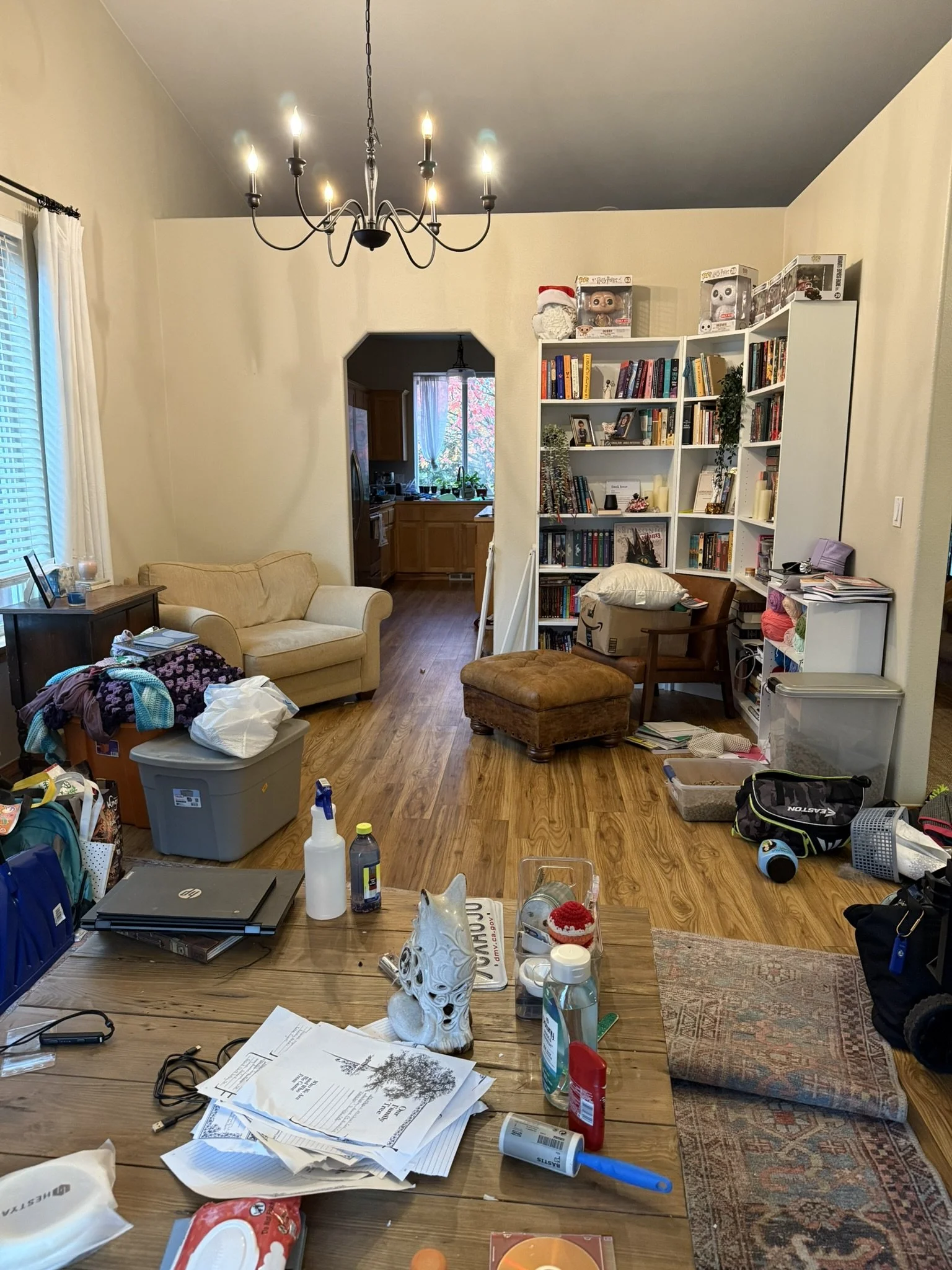 Living room with a chandelier, bookshelf, couch, and cluttered table with papers, bottles, and miscellaneous items.