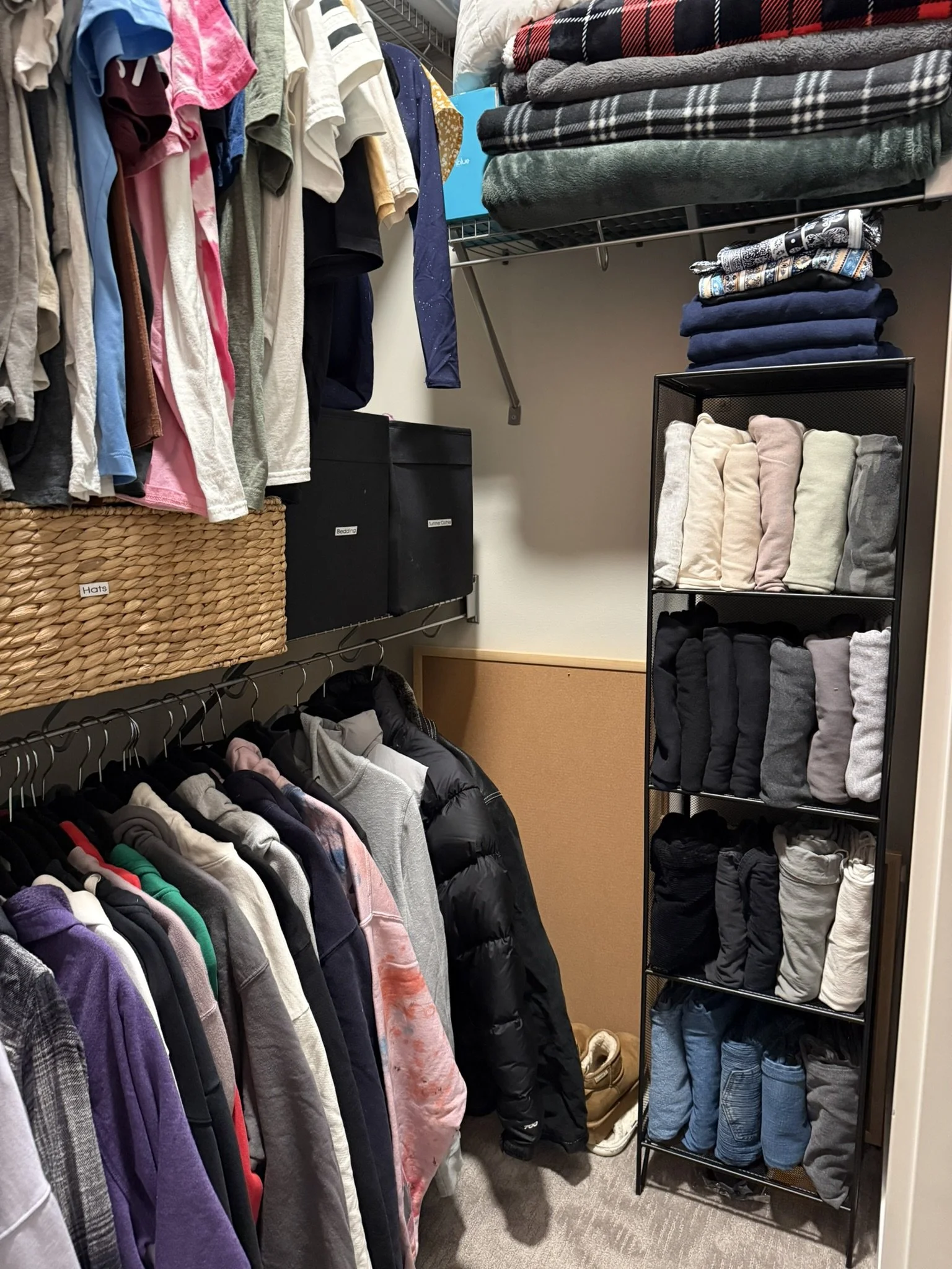 A closet with hanging shirts, folded blankets on top shelf, a black shoe rack with folded socks and clothes, and a black coat hanging among other jackets and sweatshirts.