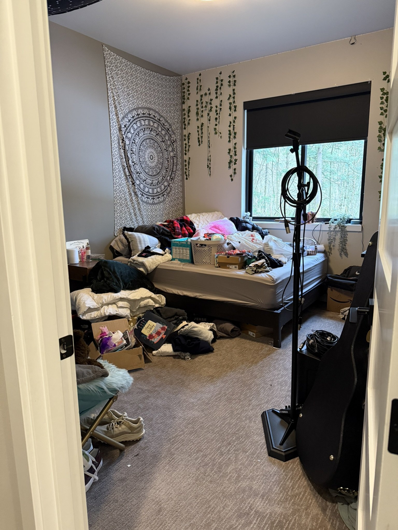 A cluttered bedroom with an unmade bed, clothes and boxes on the floor, and a window with a black blind. There is a tapestry and faux vines on the wall, and a guitar case leaning against the wall.