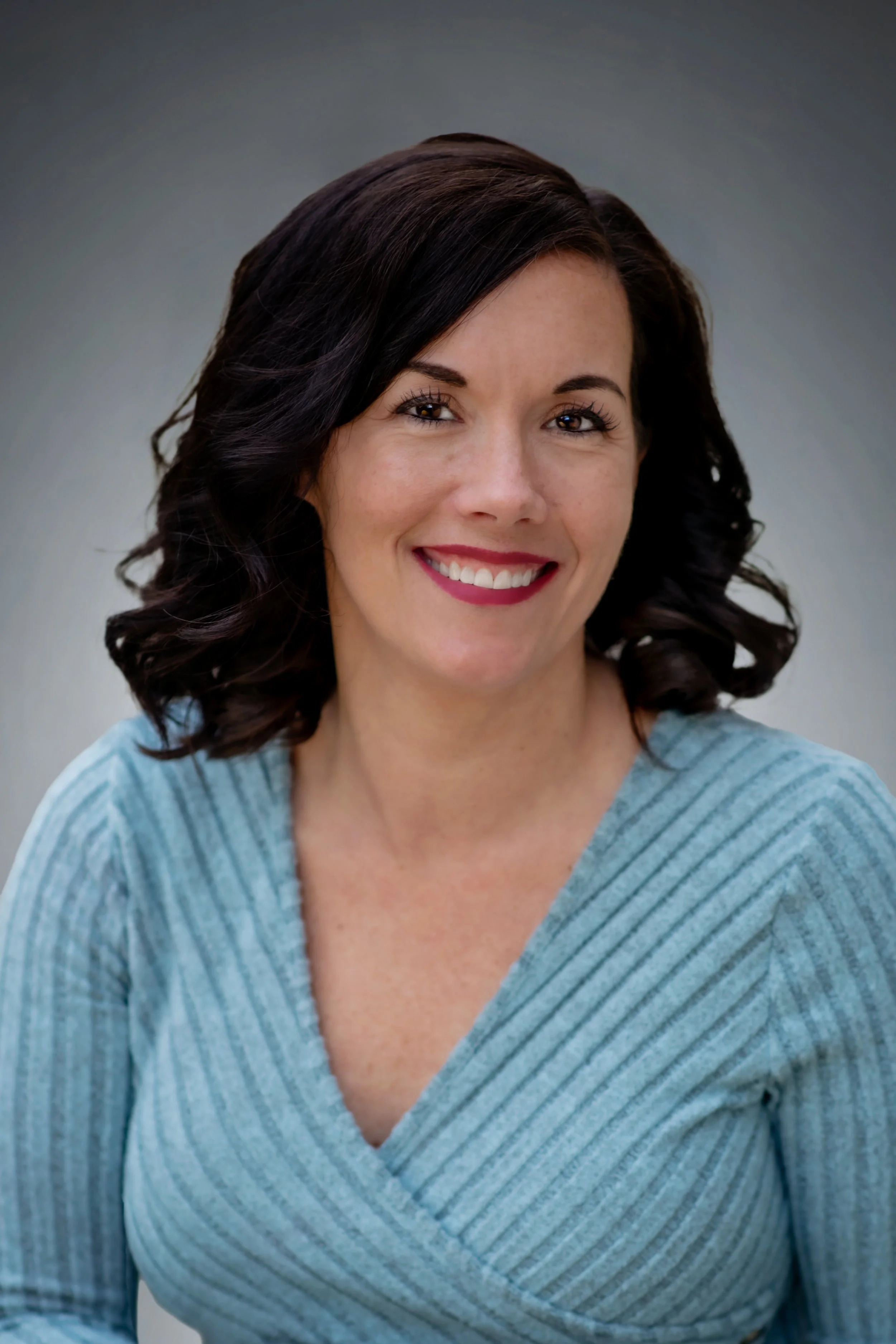 A woman with shoulder-length dark curly hair, smiling, wearing a light blue striped top against a blurred grey background.