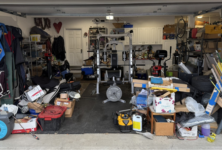 A cluttered garage filled with various tools, boxes, and fitness equipment, including a weight bench and treadmill, with shelves containing items and a white cabinet in the background.